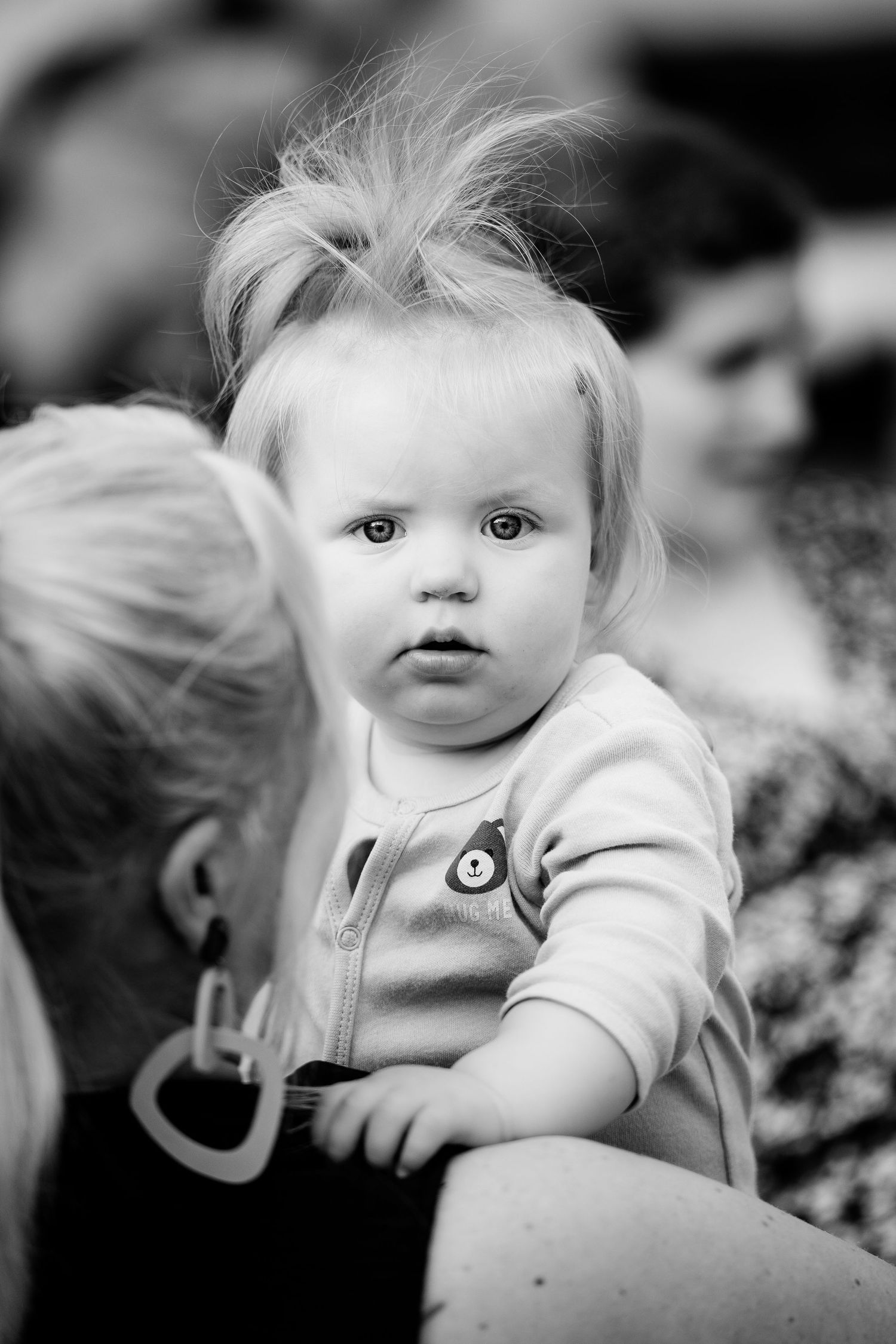 Black and white portrait of a curious baby with wispy hair sitting on an adults lap outdoors.