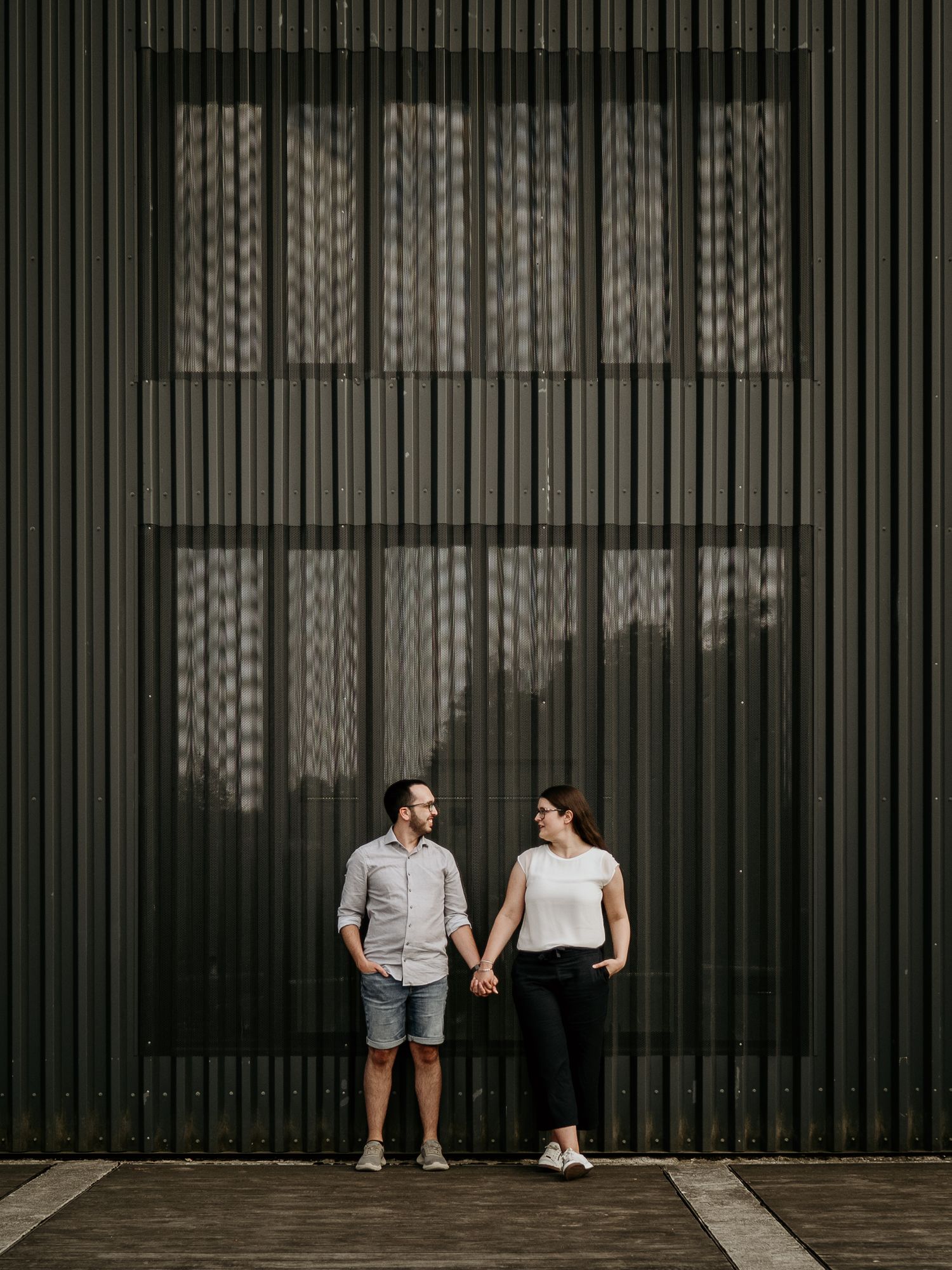 Couple holding hands and gazing at each other against a dark corrugated metal wall outdoors.