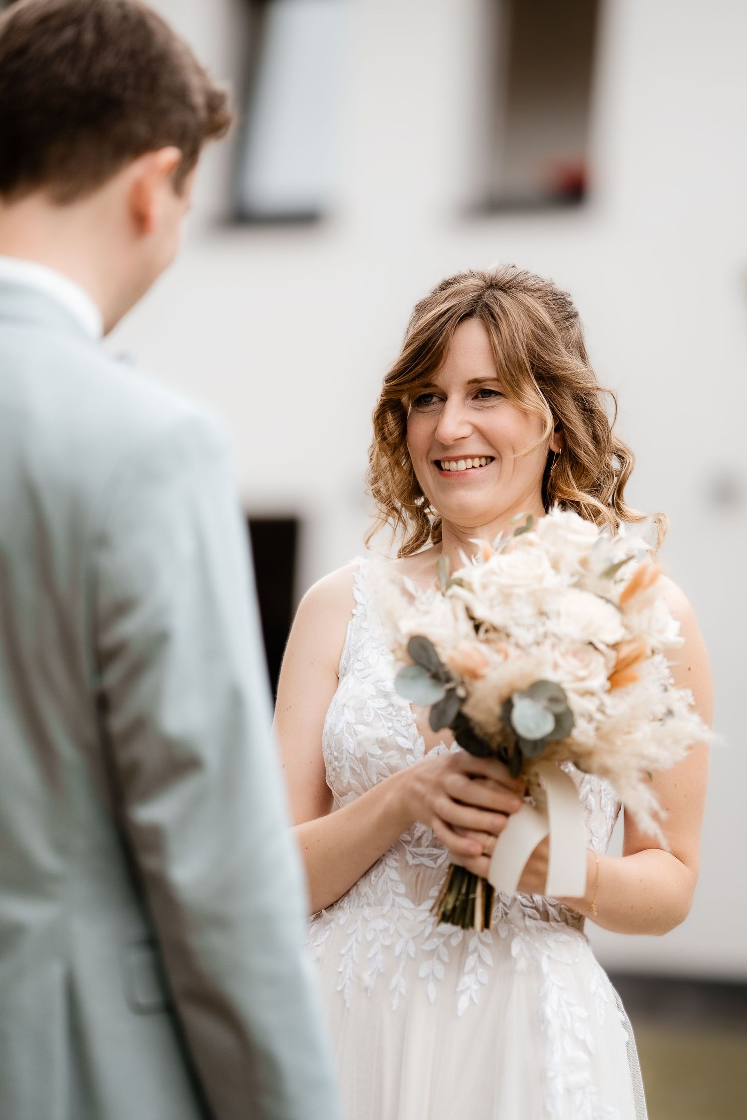 Smiling bride in lace wedding dress holds bouquet facing groom in light gray suit outdoors.