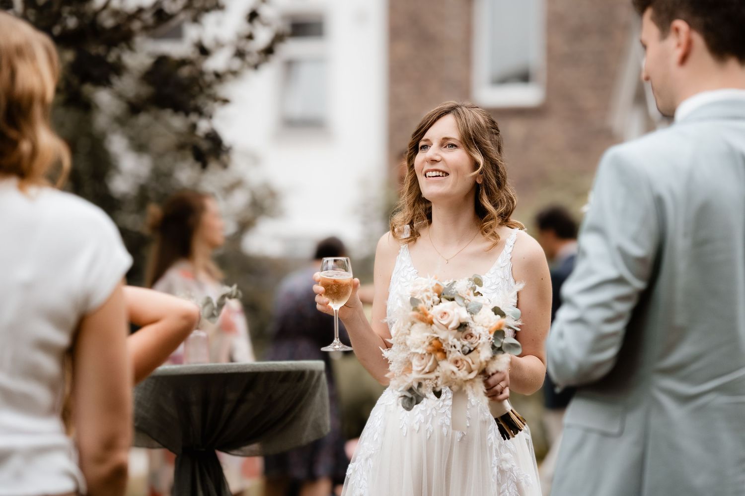 Happy bride in white lace gown holding bouquet and champagne glass at outdoor wedding reception.