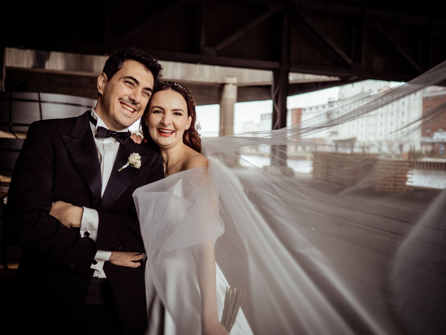Couple poses in wedding attire, bride in white off-shoulder gown with flowing veil, groom in black tuxedo, urban backdrop.