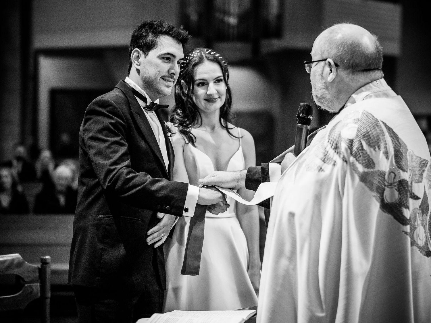 A black and white photo of a wedding ceremony with a bride, groom and priest exchanging rings at the altar.
