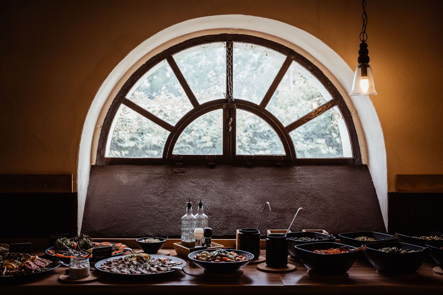 Elegant wedding reception buffet spread on wooden table beneath arched window with warm Edison bulb lighting.