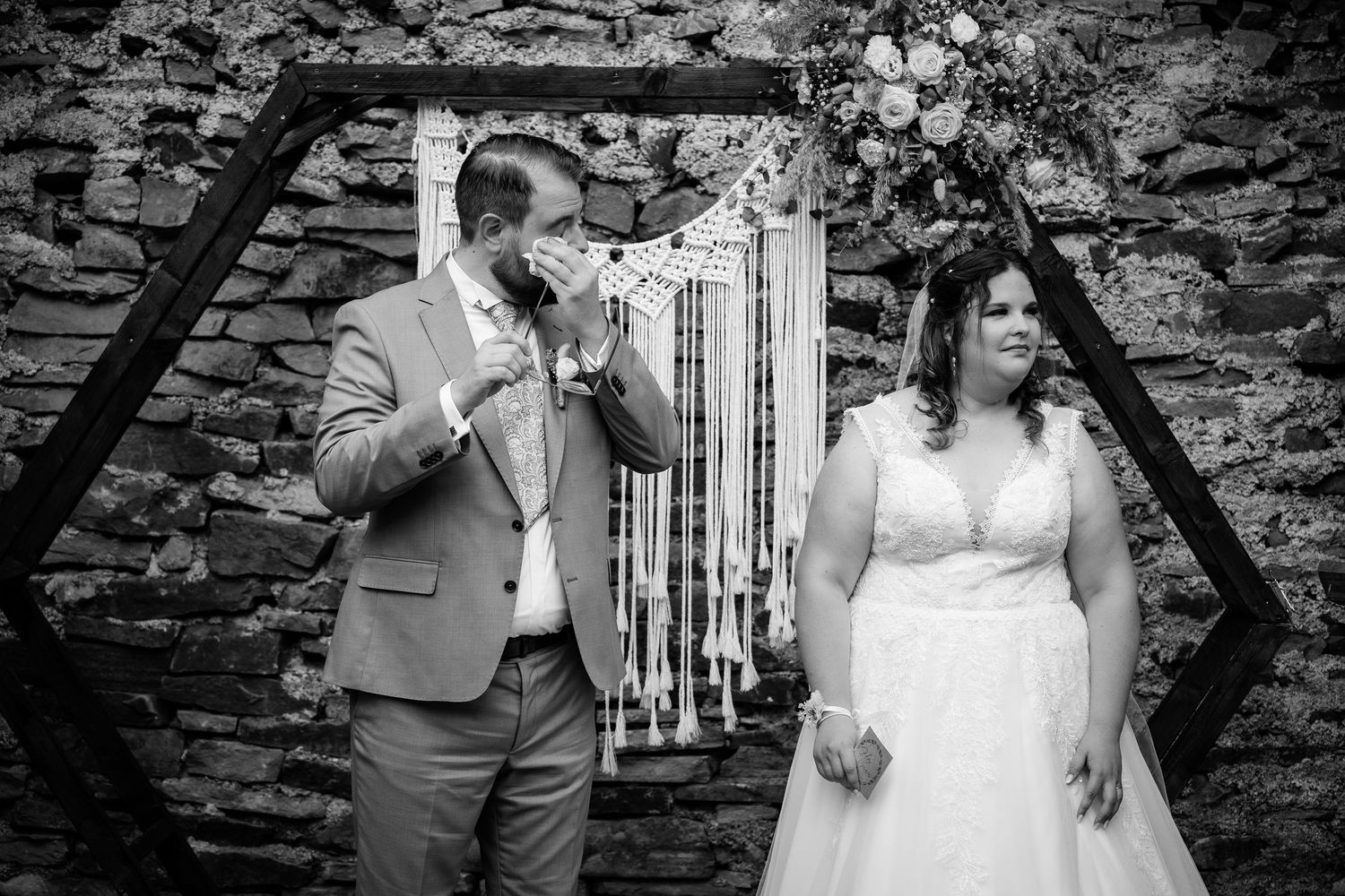 Groom wiping tears at hexagonal wedding arch with macrame backdrop beside bride in white gown.