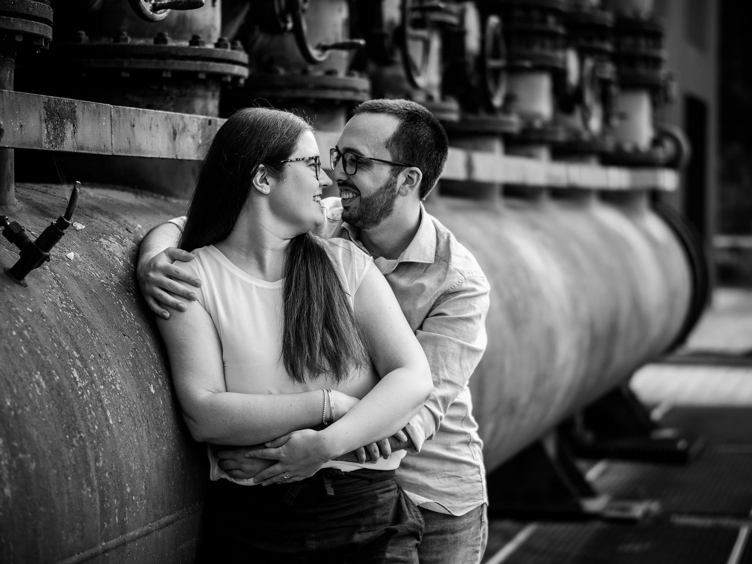 Black and white photo of a romantic couple embracing and gazing at each other near large industrial pipes.