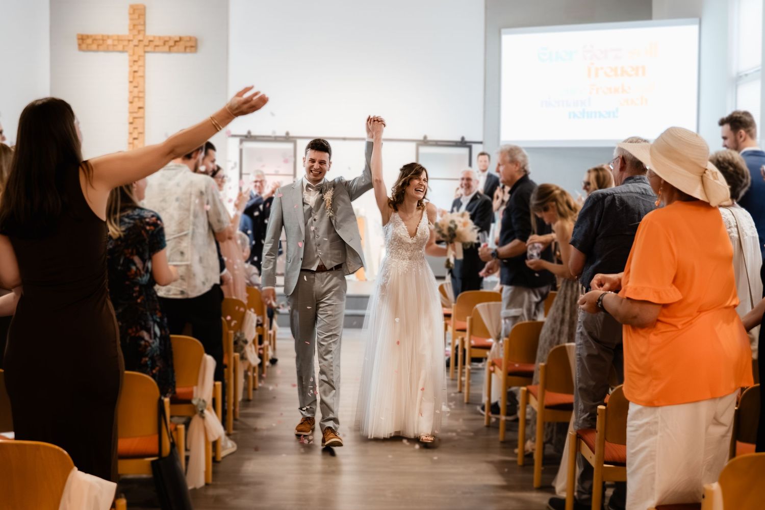 Joyful bride and groom raise hands walking down aisle after ceremony with cheering guests.