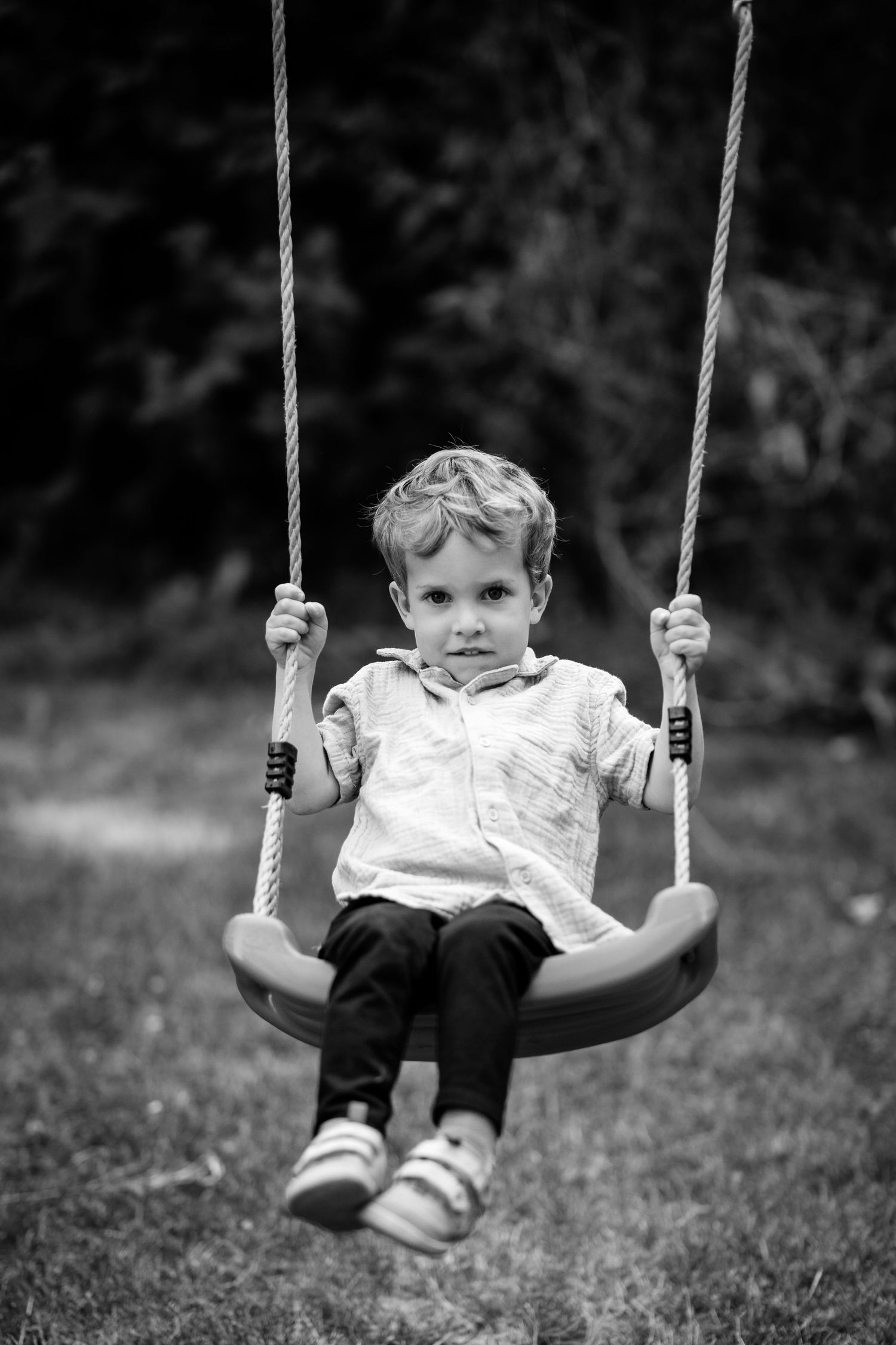 A young boy sits on a rope swing in a garden, looking at the camera in a black and white portrait photo.
