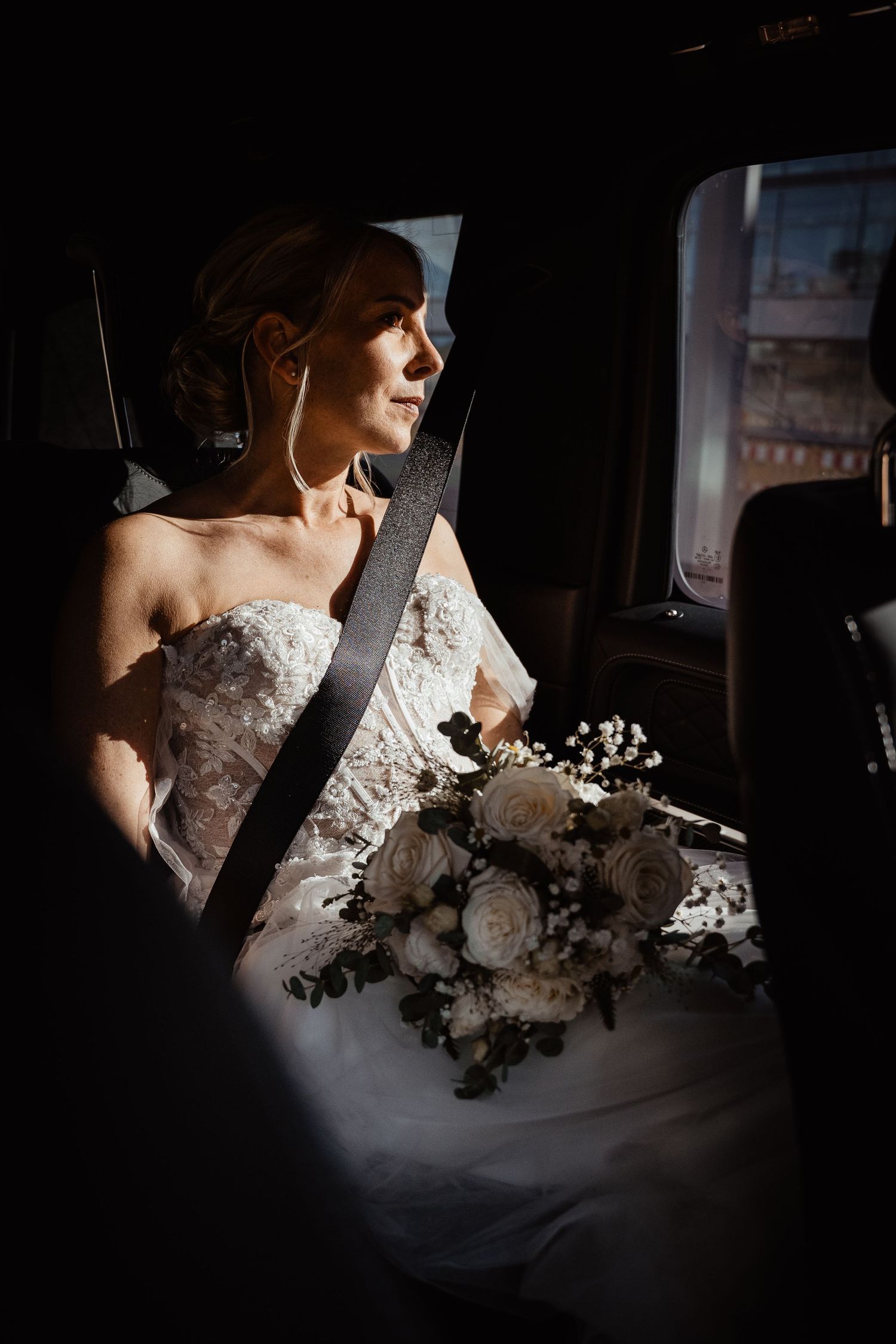 Bride in lace wedding dress holding white rose bouquet sitting in a car looking out window.