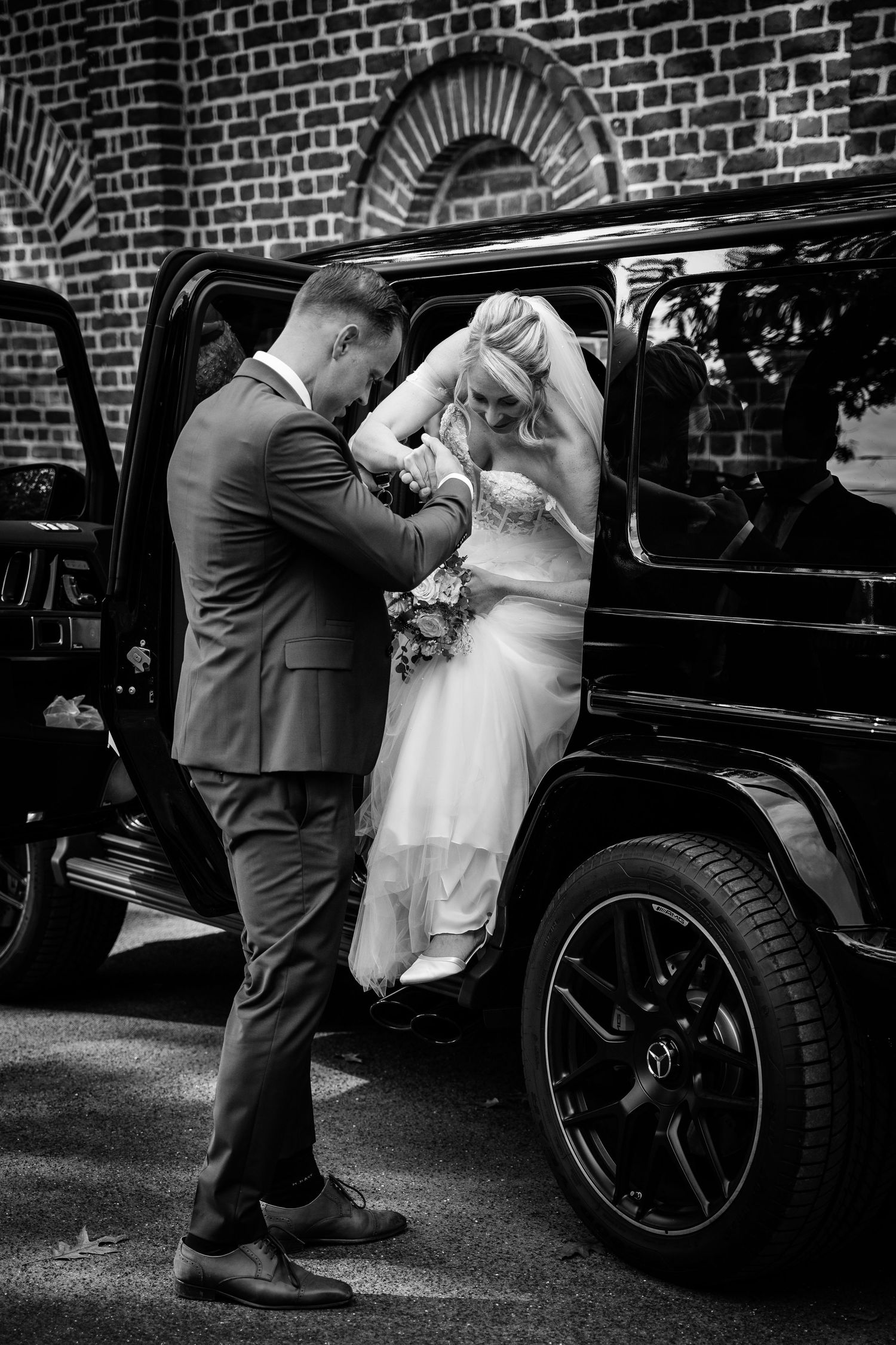 Groom helping bride exit black Mercedes G-Wagon in black and white wedding photo.