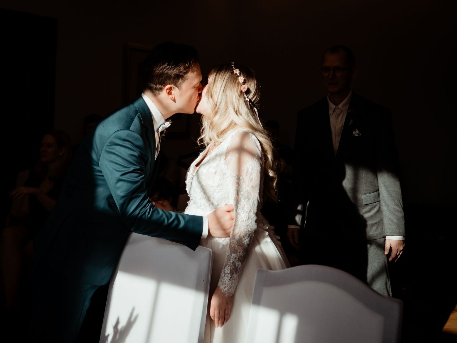A bride and groom share their first kiss at a wedding ceremony, bathed in dramatic natural light and shadows.