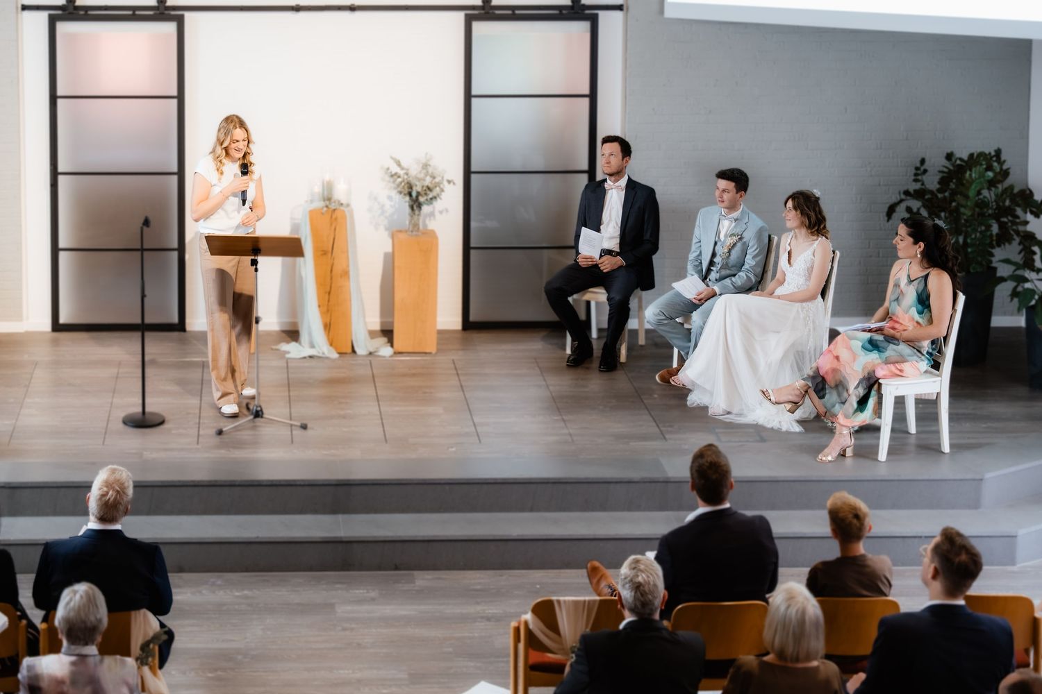 A woman speaks at a podium during an elegant indoor wedding ceremony while the couple and guests look on.