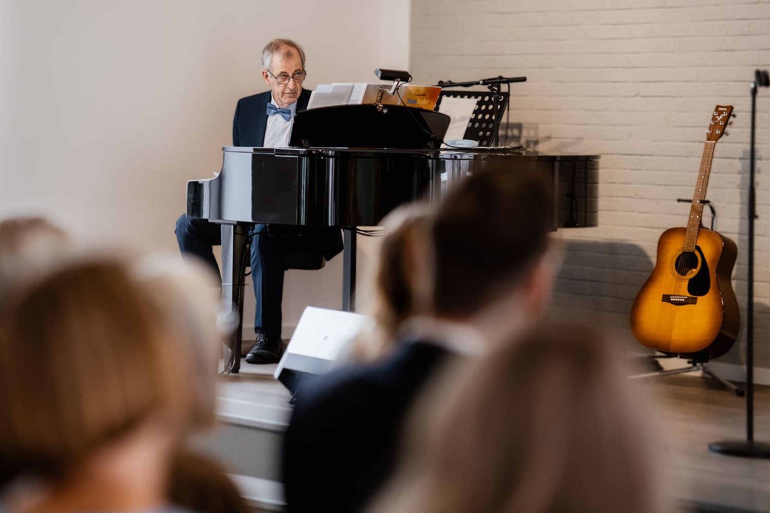Man playing grand piano at intimate recital with acoustic guitar and seated audience in bright modern room.