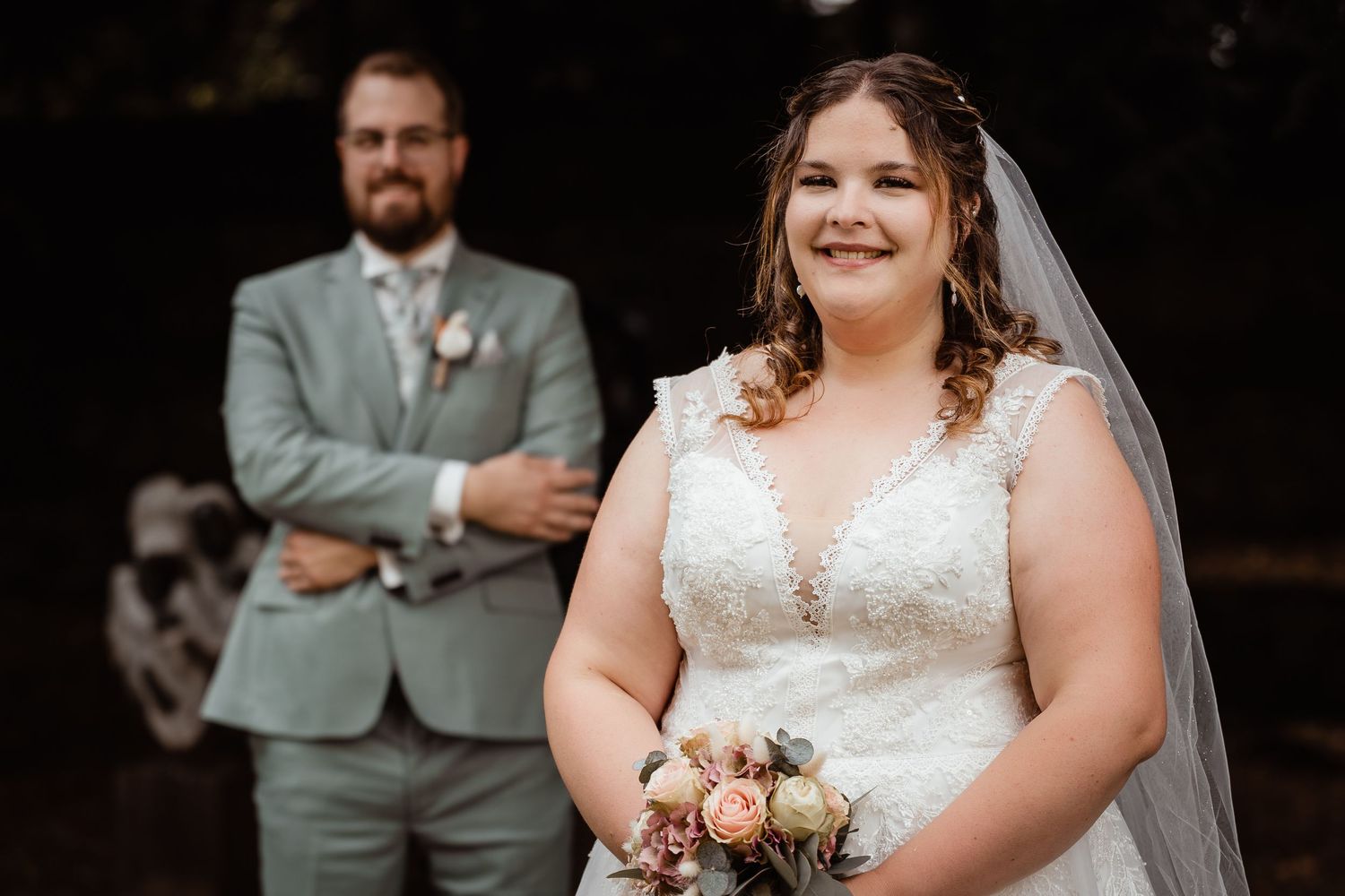 Bride in lace wedding dress holding floral bouquet with groom in grey suit standing behind her.