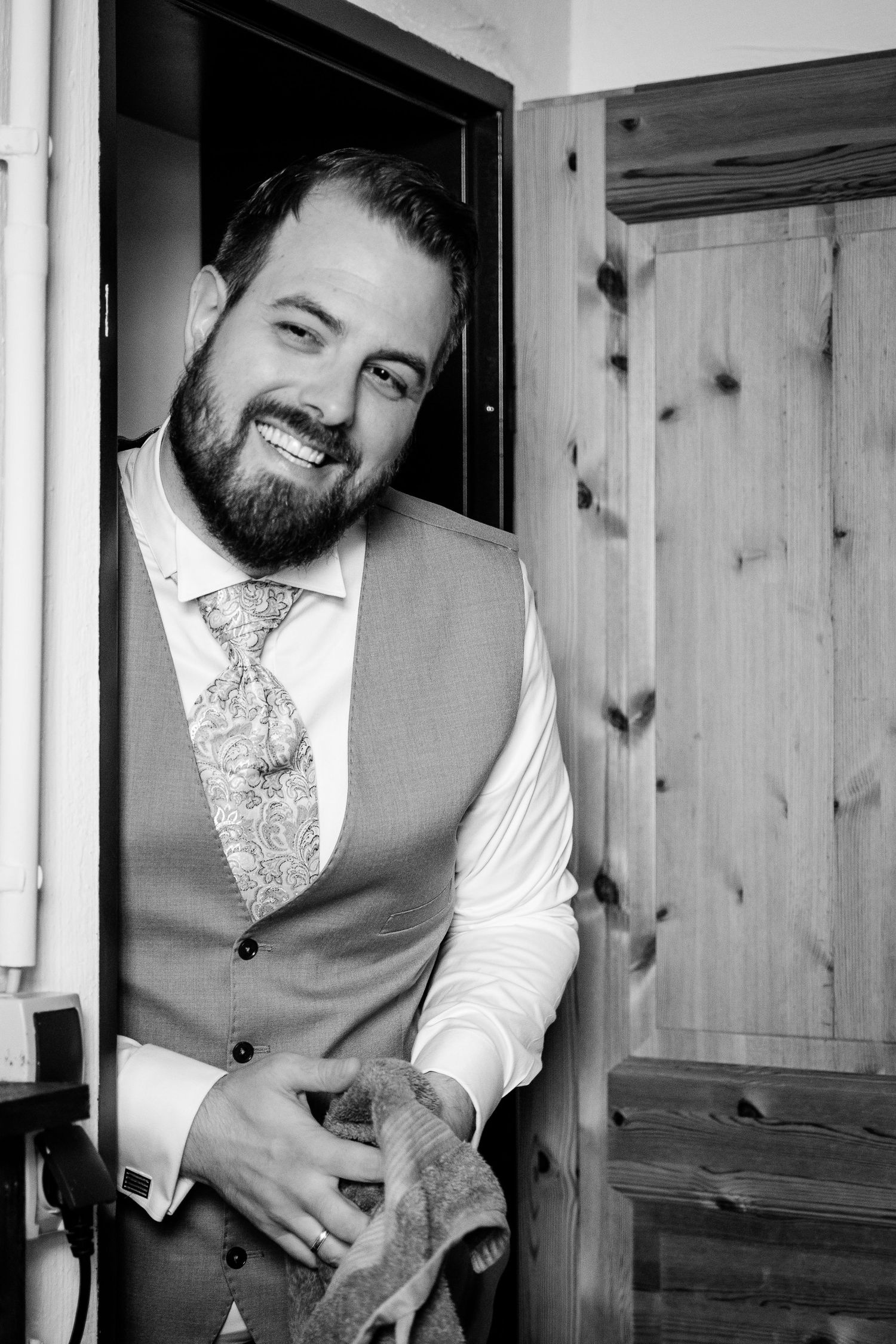 Smiling bearded groom in vest and paisley tie leaning against wooden door in black and white portrait.