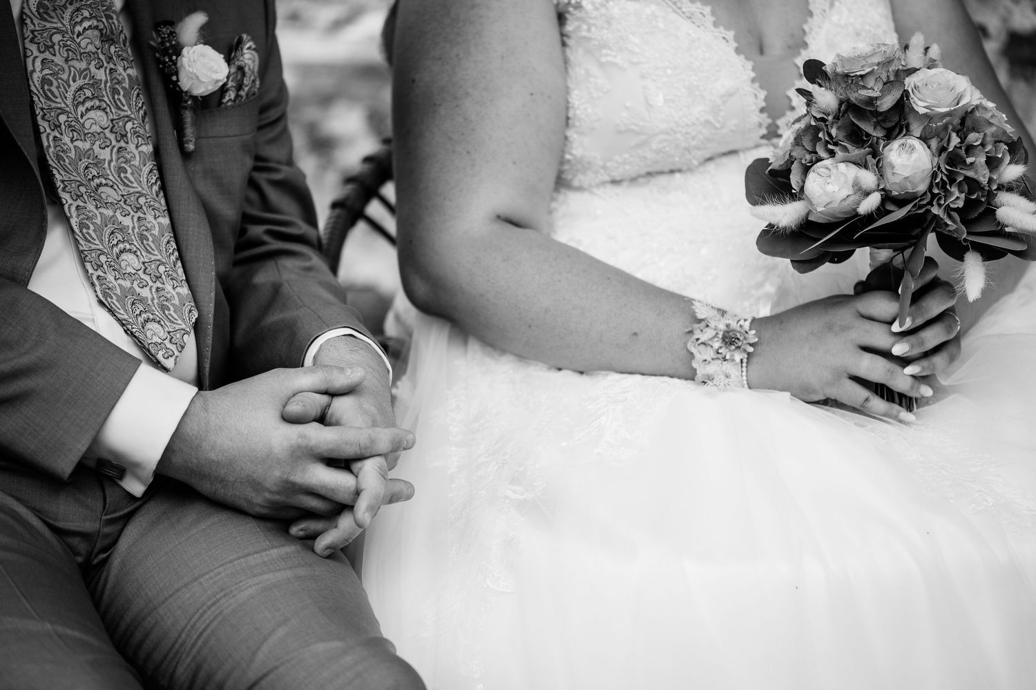 Black and white photo of bride and groom holding hands during wedding ceremony, bride holding floral bouquet.