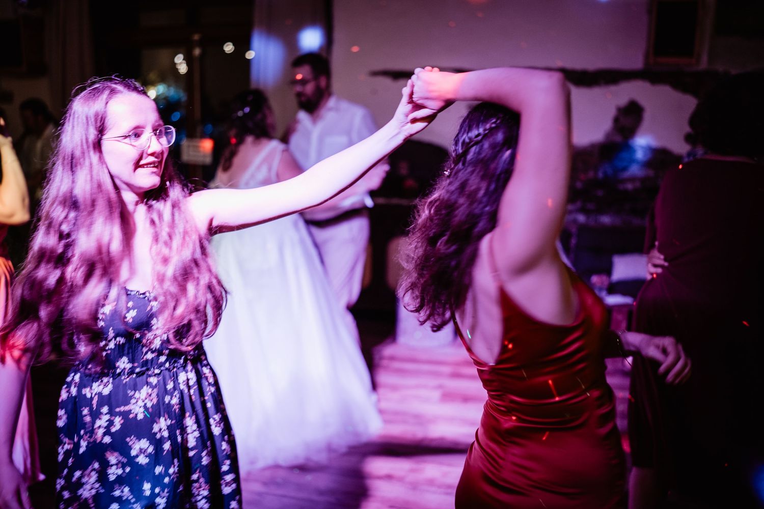 Two women dancing together joyfully at wedding reception under colorful party lights.