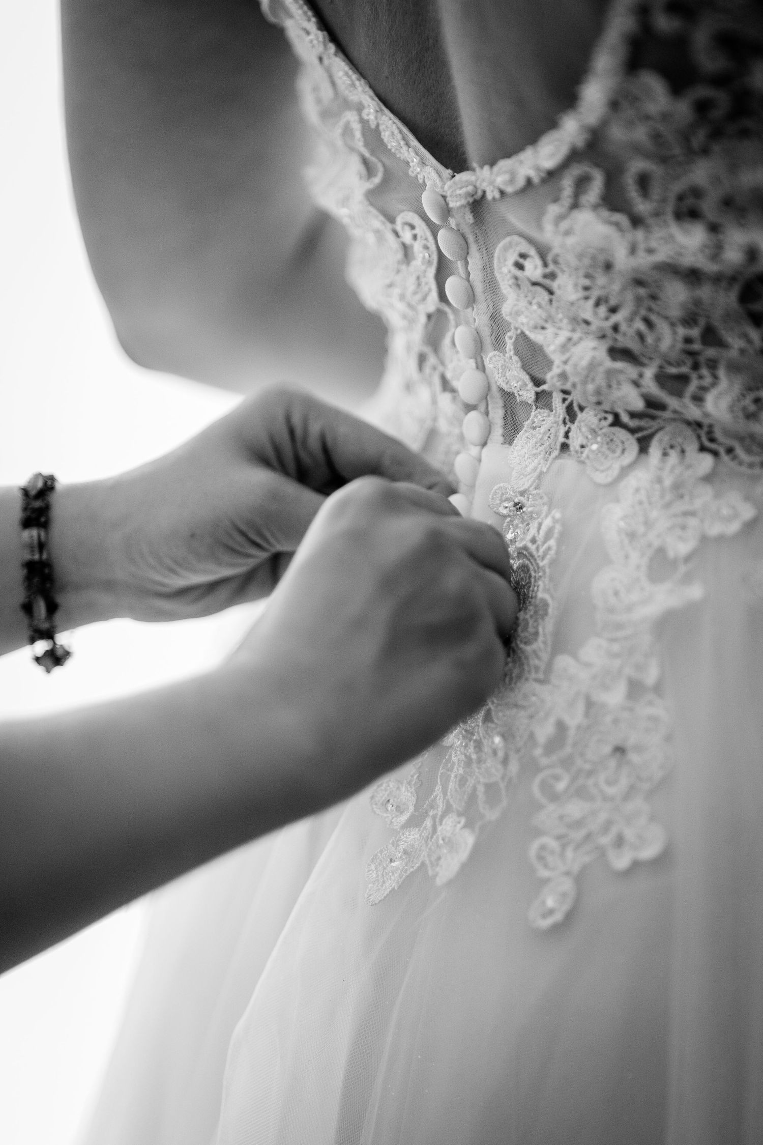 Black and white photo of hands buttoning the back of a lace wedding dress on a bride.