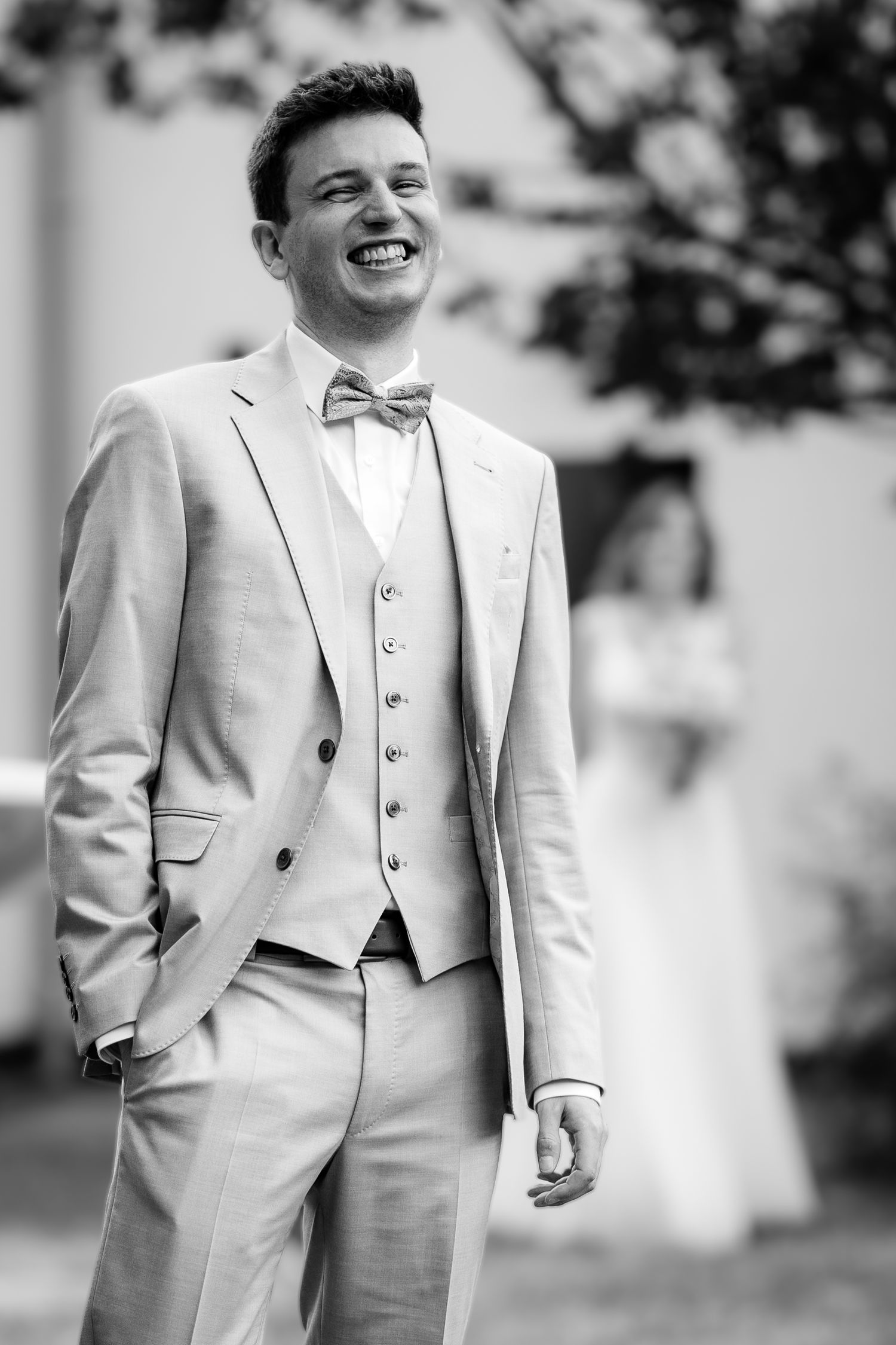 Black and white photo of a smiling groom in a light suit and bow tie at a wedding ceremony.