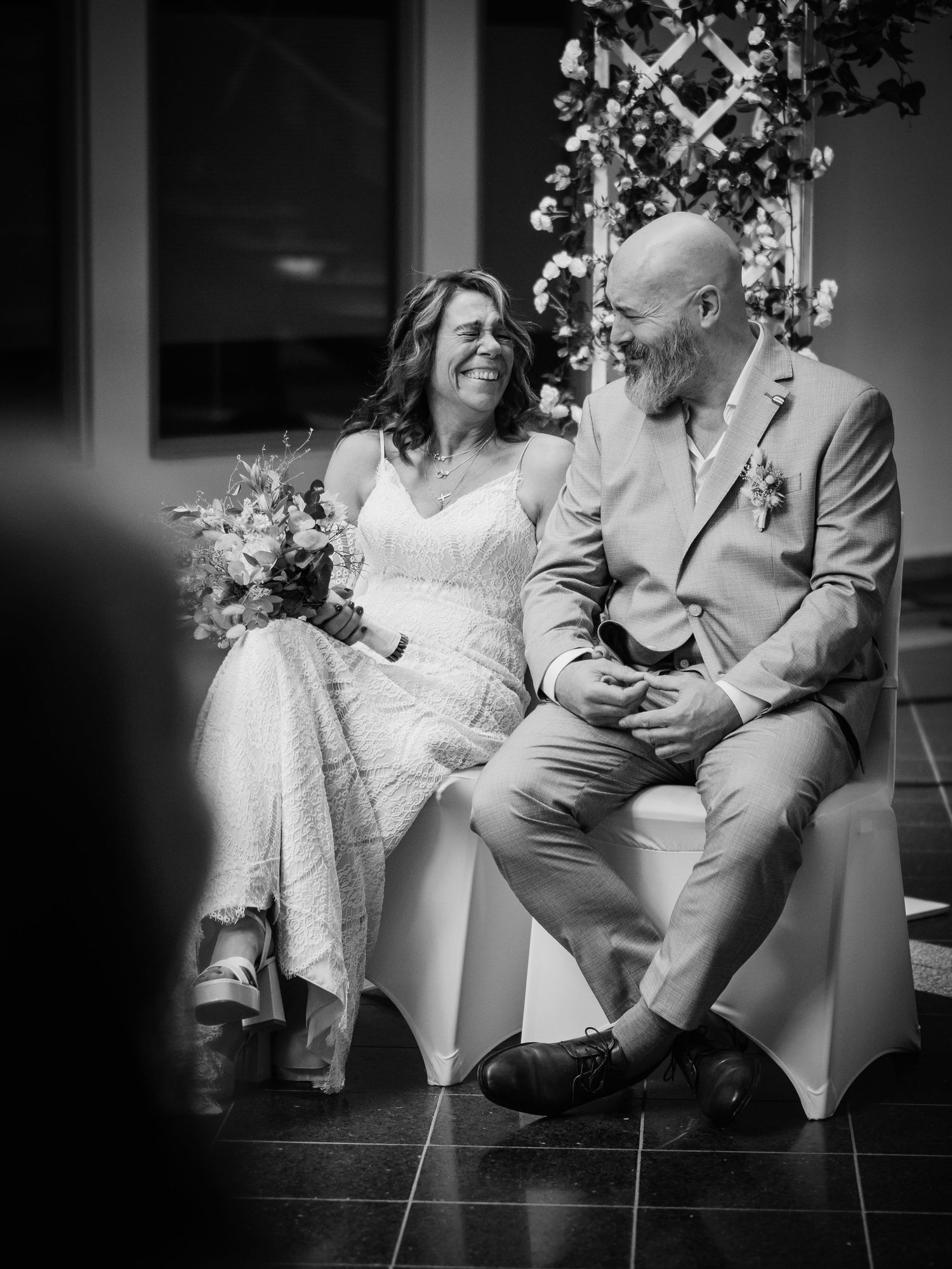 Black and white photo of a laughing bride and groom sitting together during their wedding ceremony.
