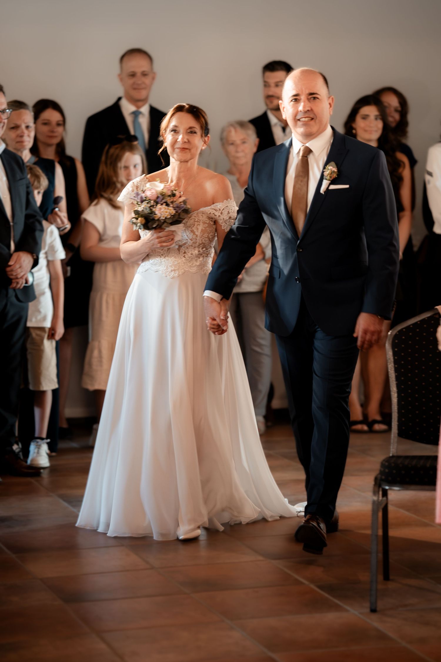 Bride and groom walk down the aisle hand in hand, smiling, with wedding guests standing in the background.
