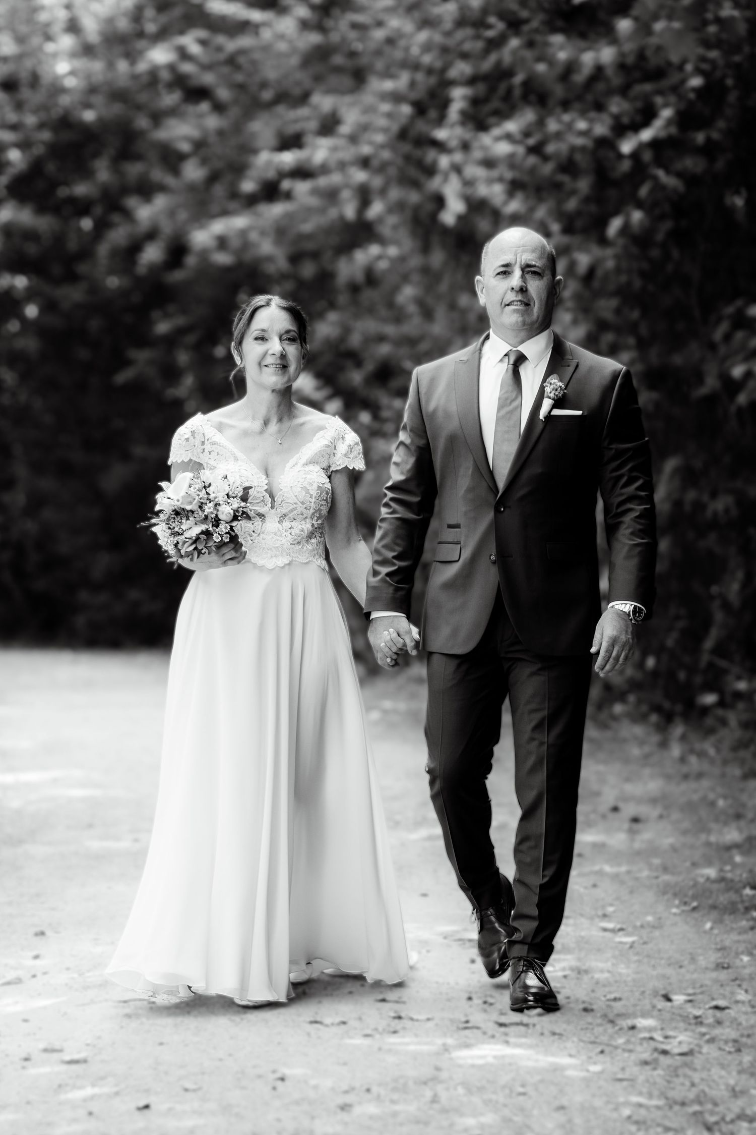 Black and white photo of bride and groom walking hand in hand on a tree-lined path on their wedding day.