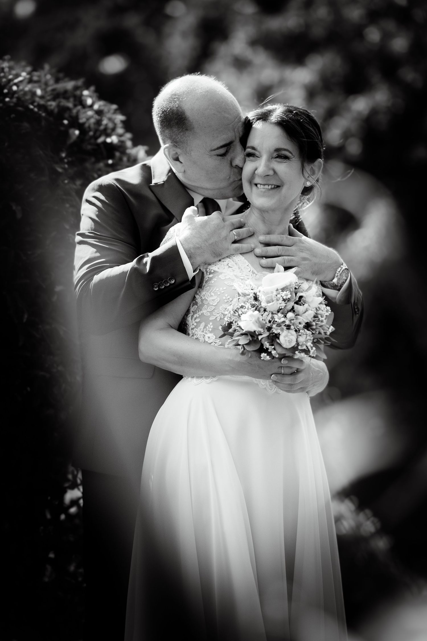 Black and white romantic portrait of groom embracing bride holding bouquet in garden setting.