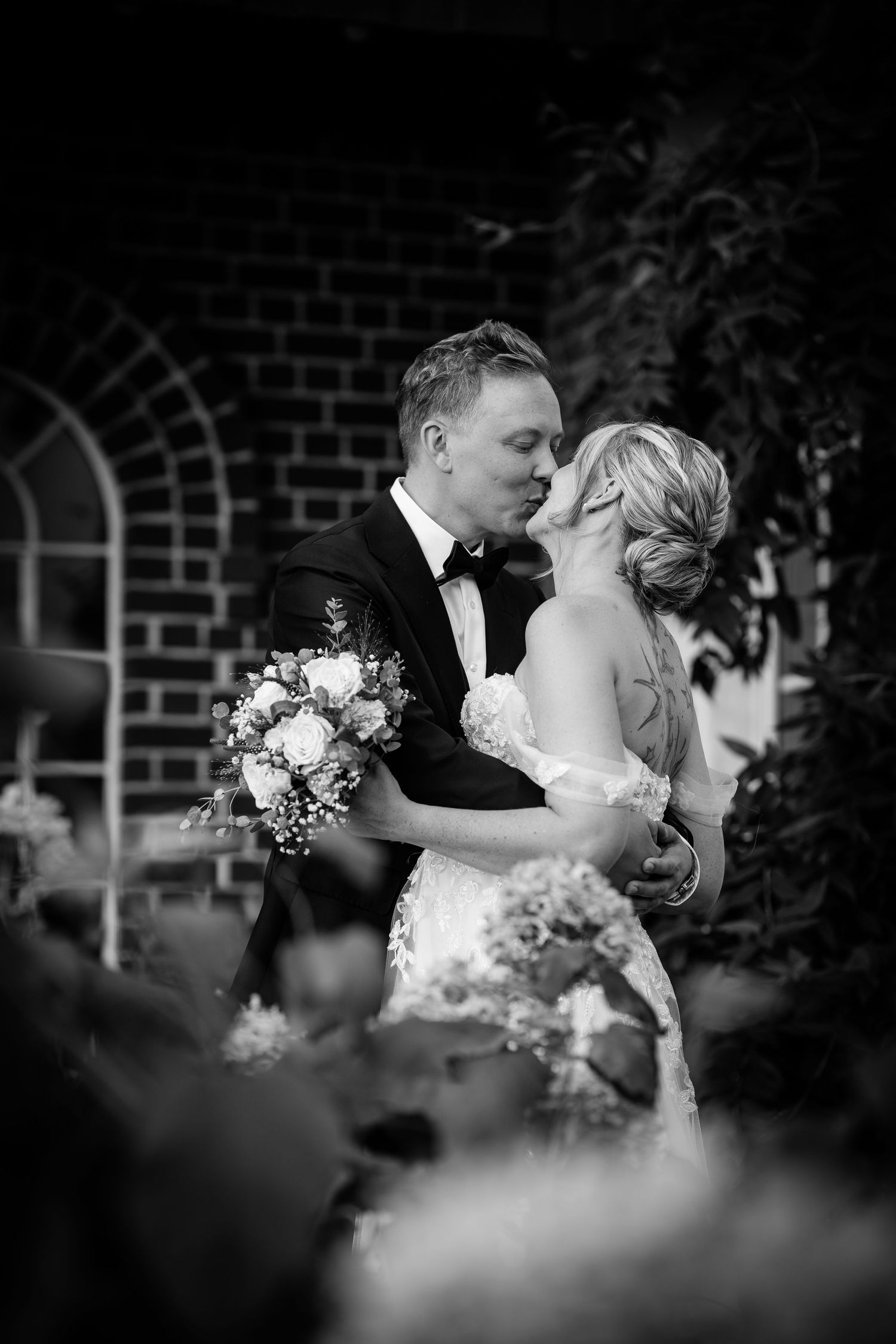 Bride and groom sharing a tender kiss outdoors surrounded by lush greenery in black and white.