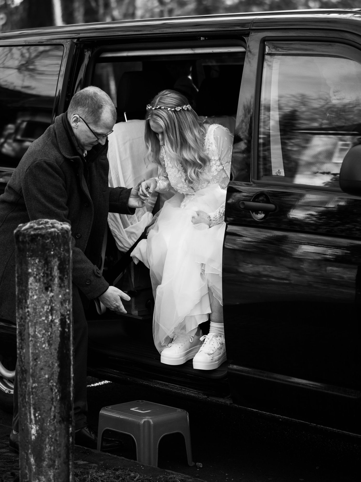 Bride in white dress and sneakers stepping out of black car, assisted by man, black and white wedding photo.