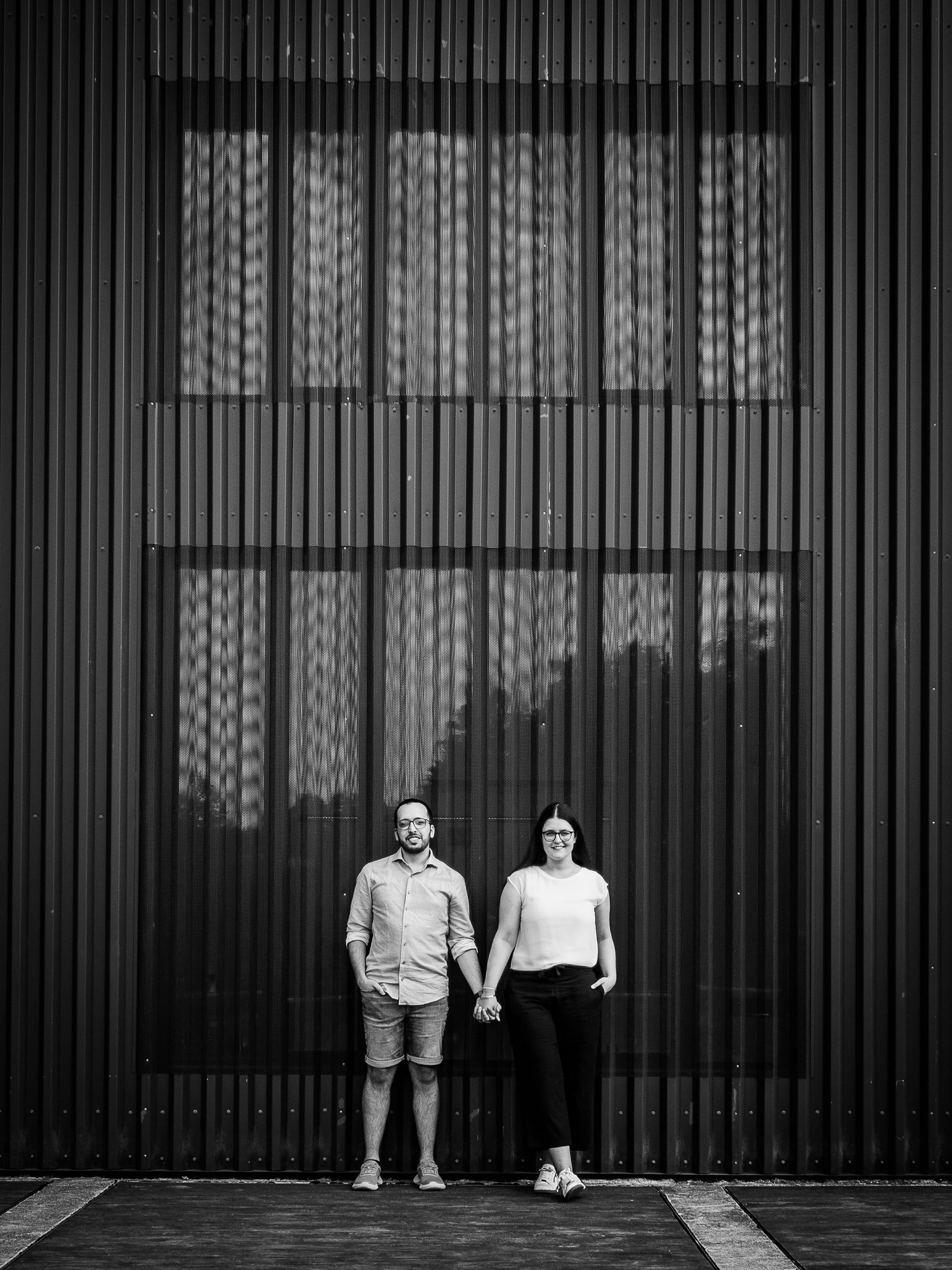 Black and white photo of a couple holding hands standing against a modern corrugated metal wall exterior.
