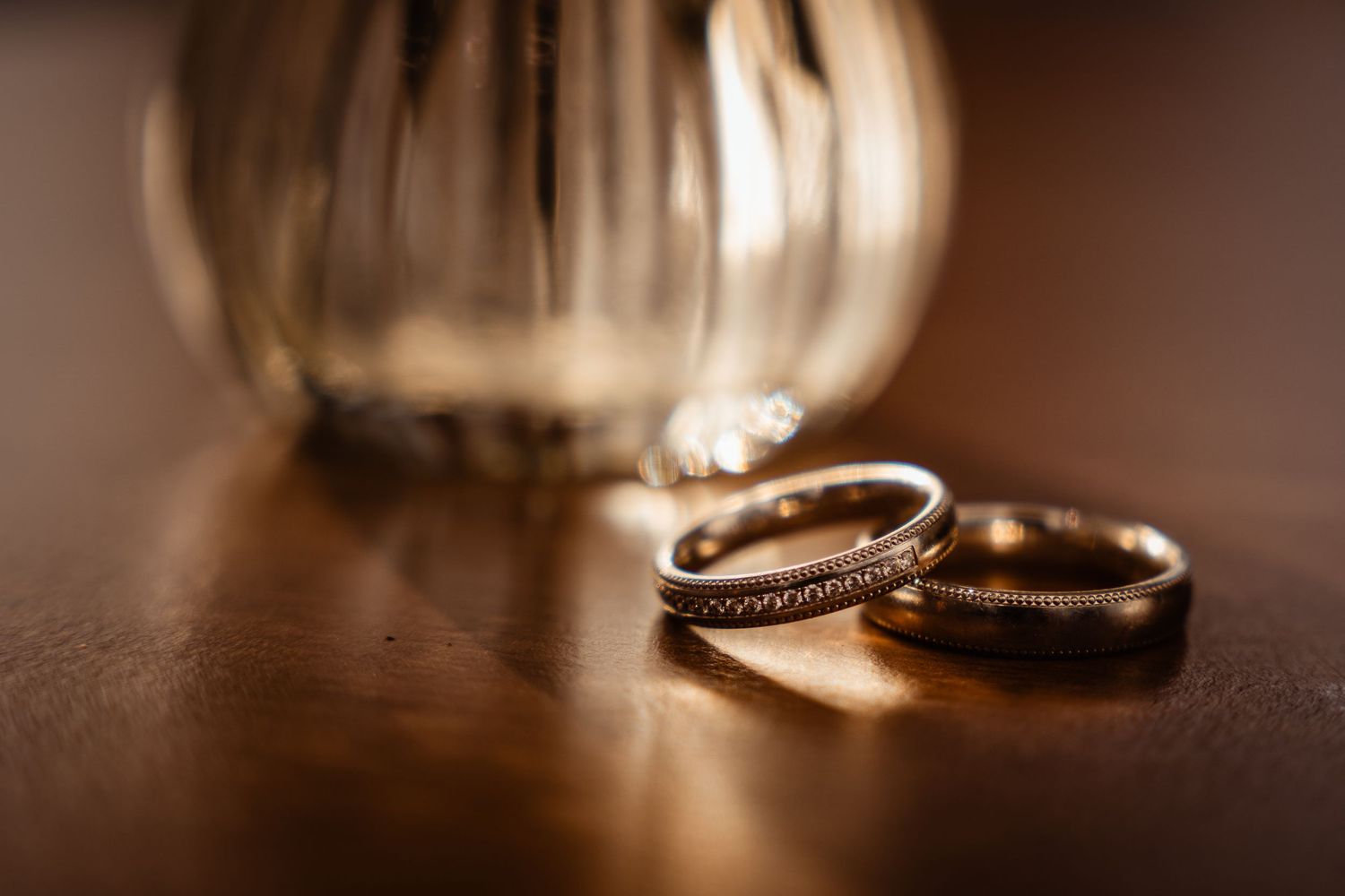 Two gold wedding bands rest on a wooden surface beside a glass vase, glowing in warm light.