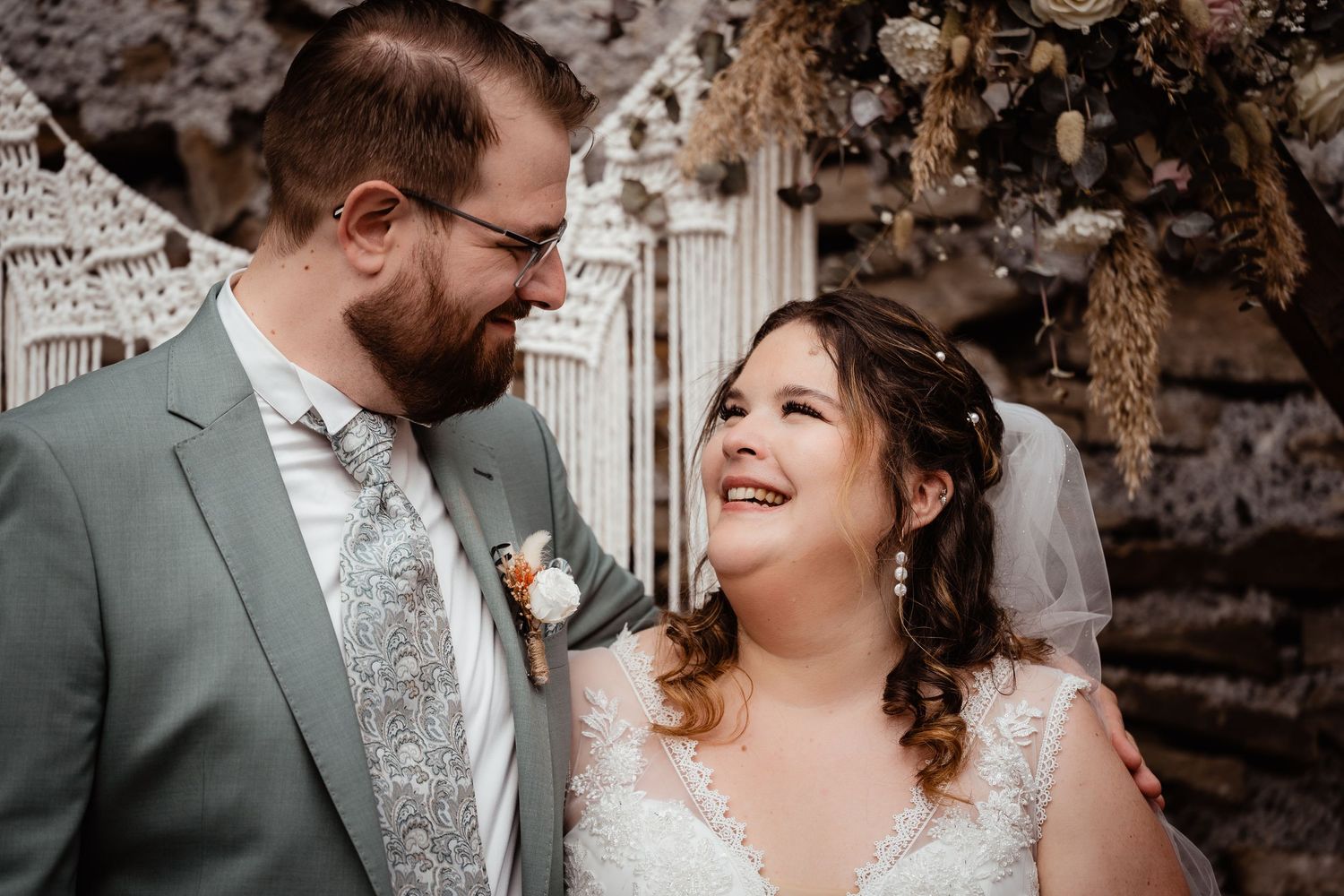 Smiling bride and bearded groom sharing a joyful moment before a rustic macrame floral wedding arch.
