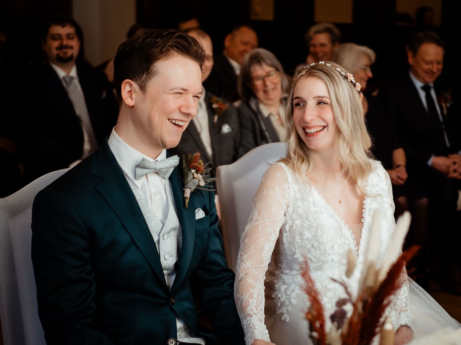 Happy couple laughing during wedding ceremony, groom in teal suit, bride in lace dress with floral headpiece.