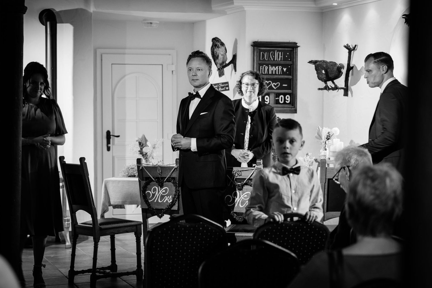 Black and white wedding reception photo showing guests and a man in a tuxedo giving a speech indoors.
