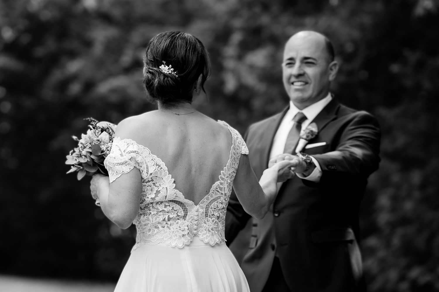 Black and white photo of a bride in lace gown holding bouquet, groom smiling and reaching out during wedding ceremony.
