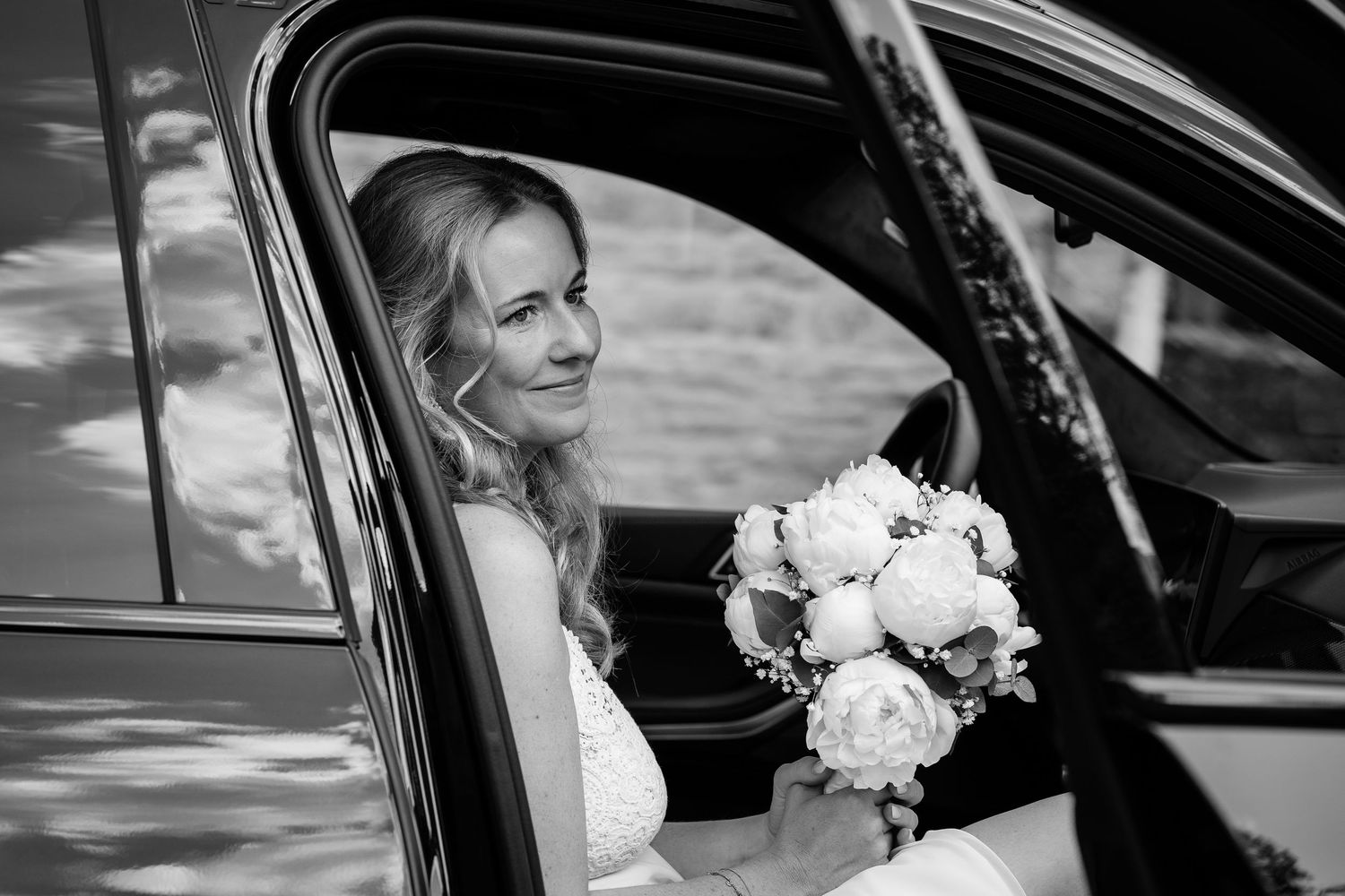 Bride in wedding dress holding white peony bouquet, seated in car window, elegant black and white portrait photo.