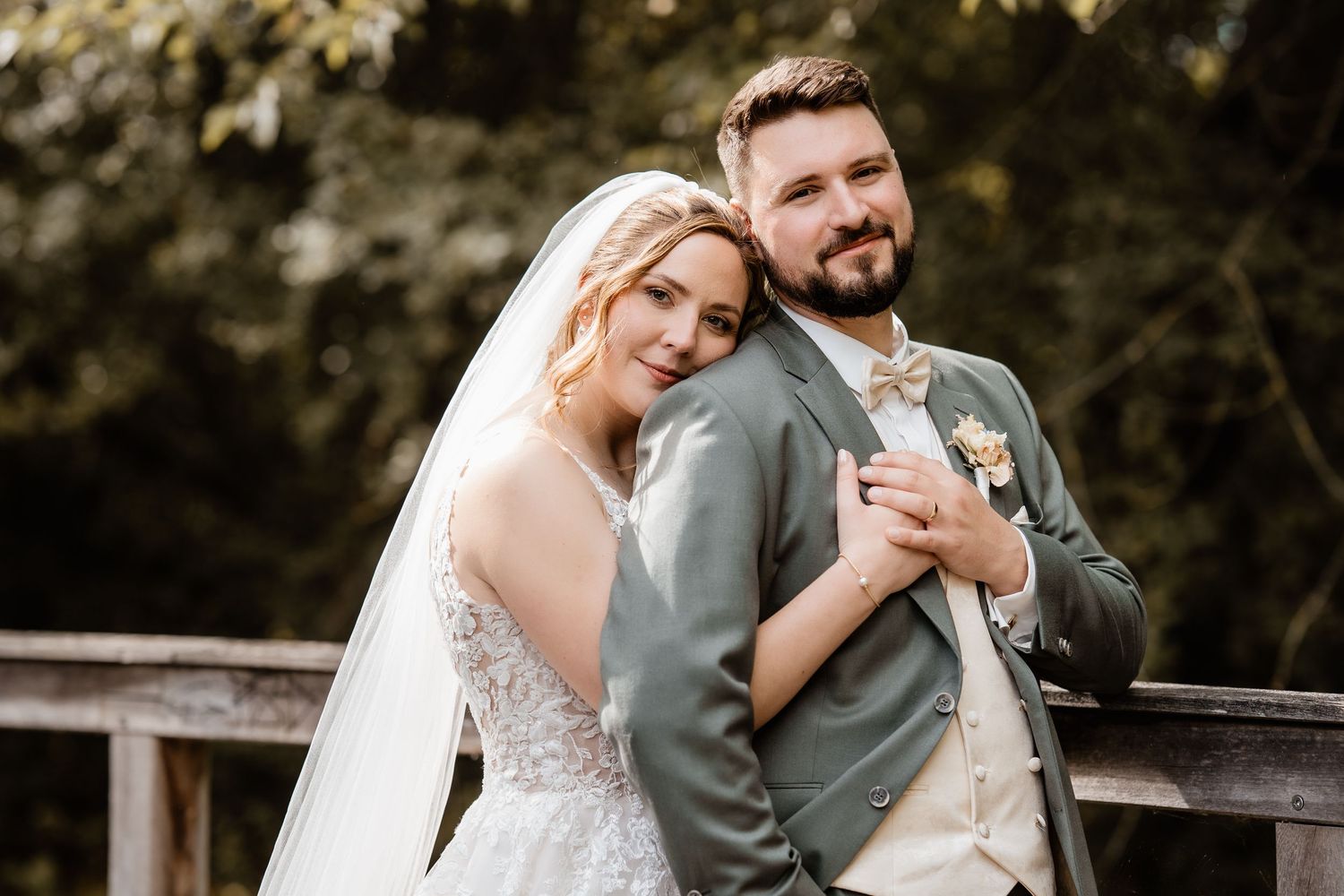 Bride in lace gown and veil leans on groom in sage suit with boutonniere on wooden bridge outdoors.