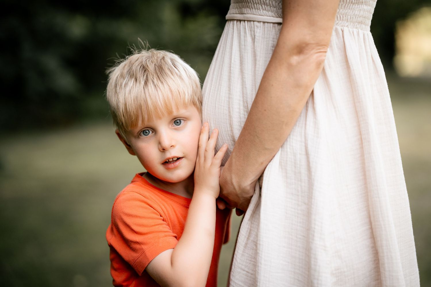 Blonde boy in orange shirt hugging pregnant mother in white dress outdoors in green park setting.