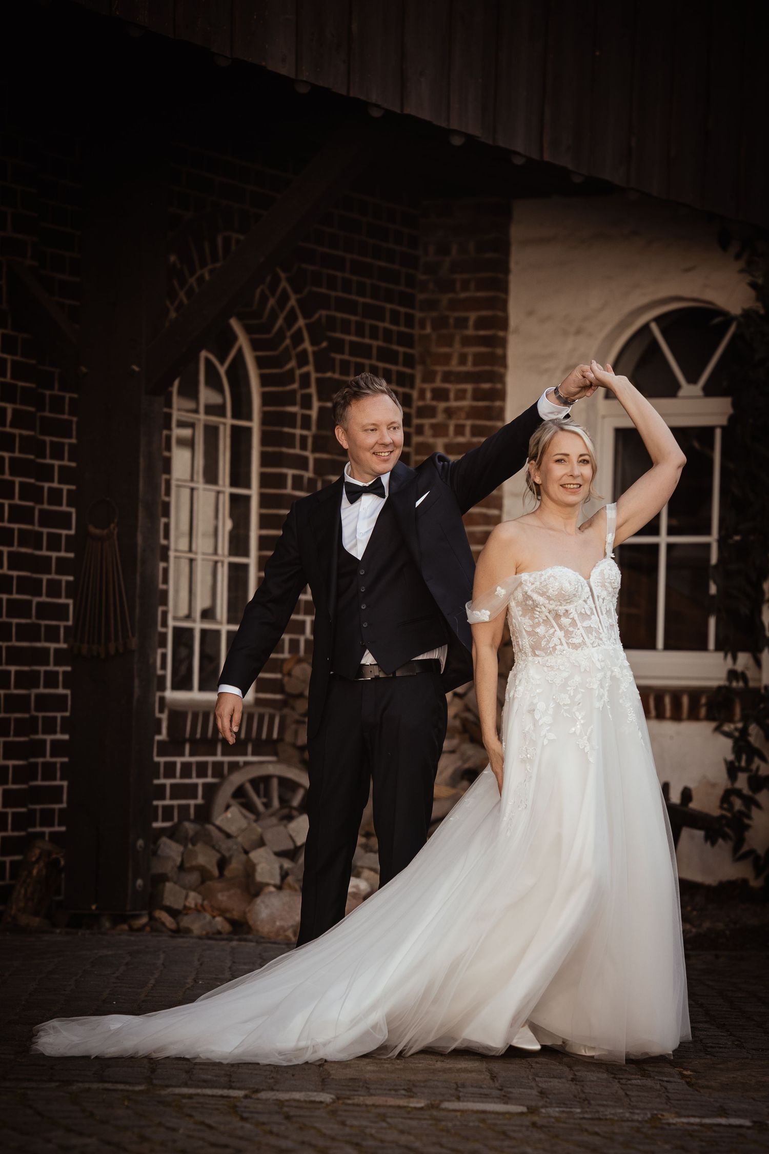 Bride and groom dancing joyfully outside a rustic brick building during their wedding celebration.