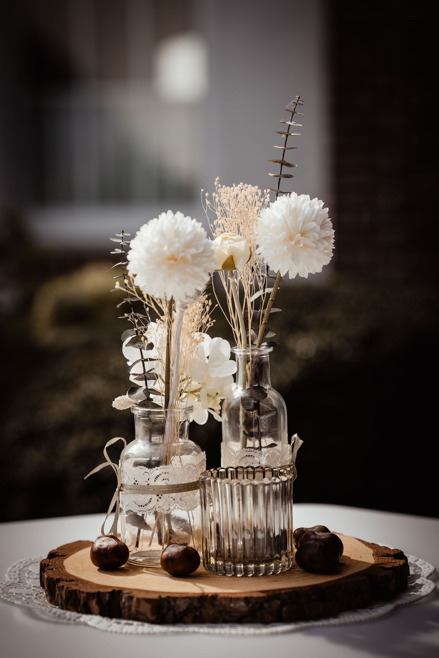 Rustic wedding centerpiece with white dried flowers in glass bottles on a wooden slice with chestnuts.