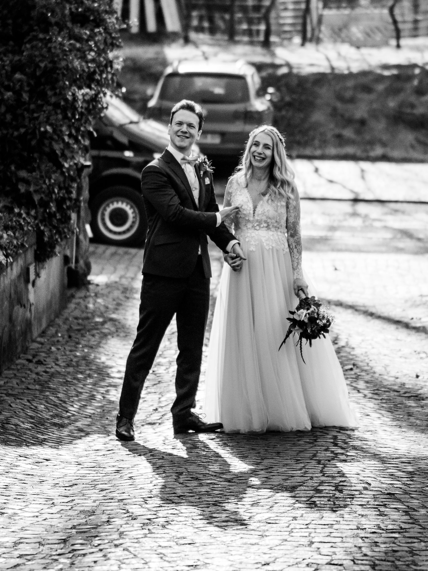 Black and white photo of smiling bride and groom holding hands on cobblestone street on their wedding day.