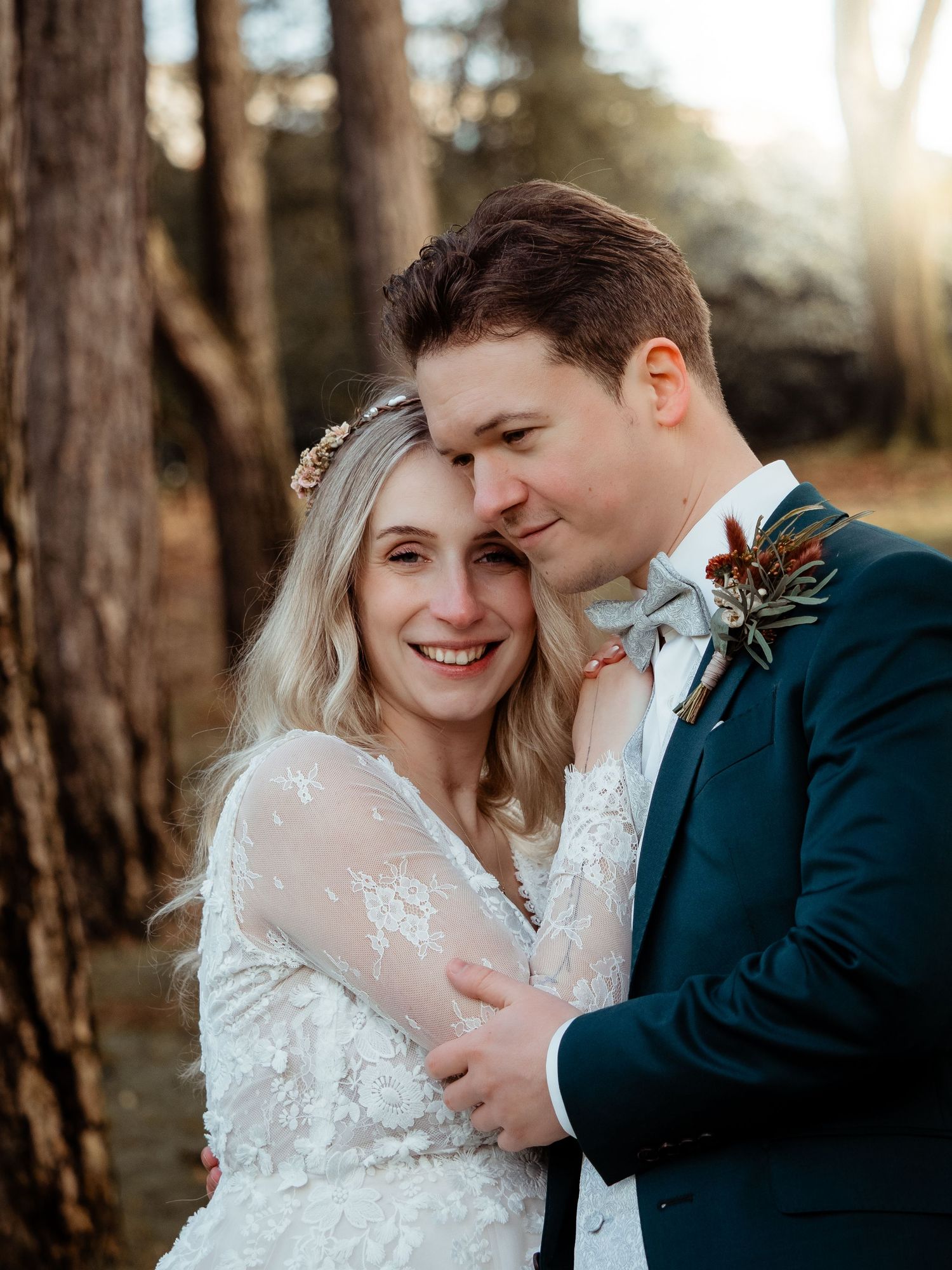 Smiling bride and groom embrace in a sun-drenched forest, groom wearing teal suit.