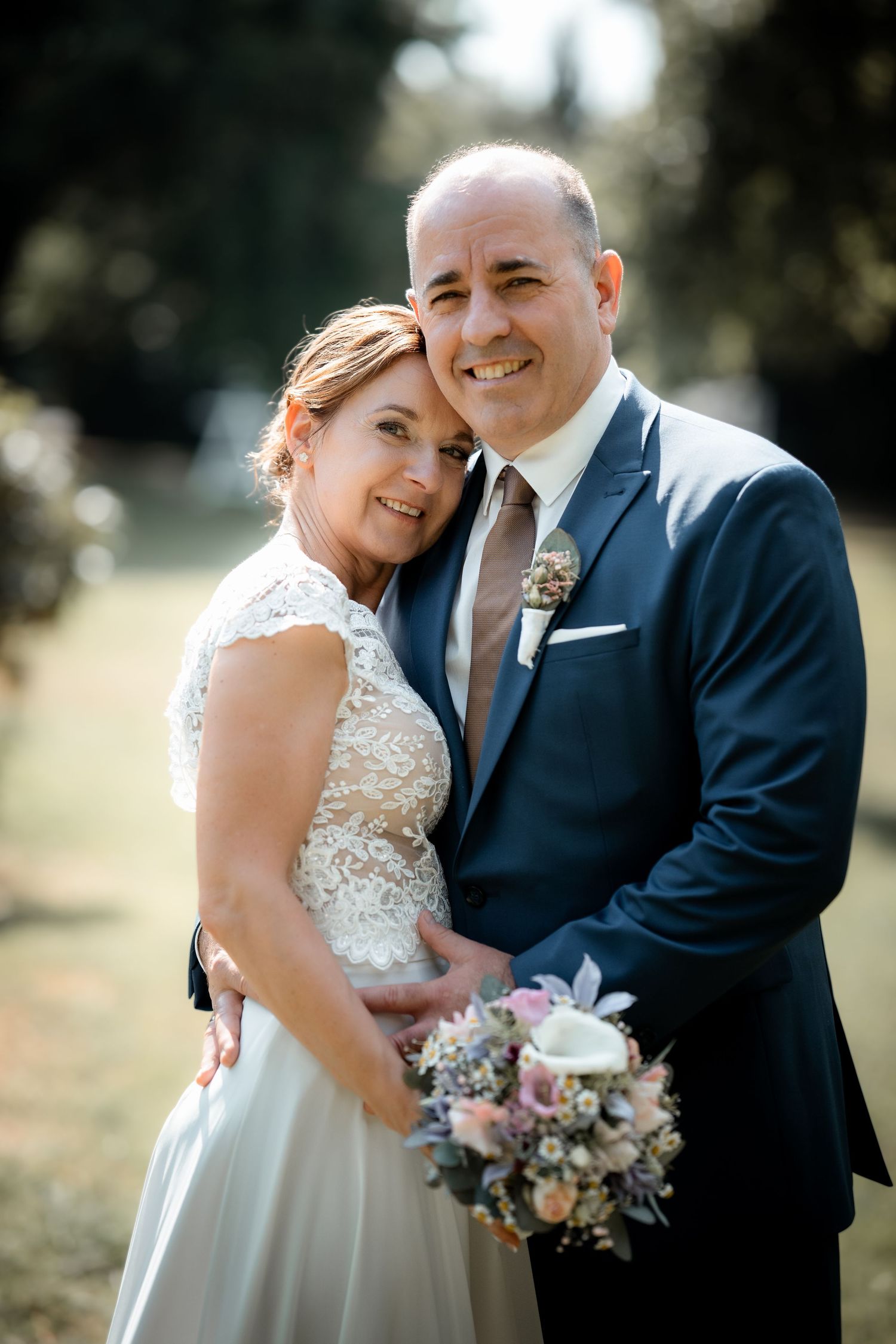 Smiling bride and groom pose closely together outdoors, holding a pastel bouquet in warm sunlight.