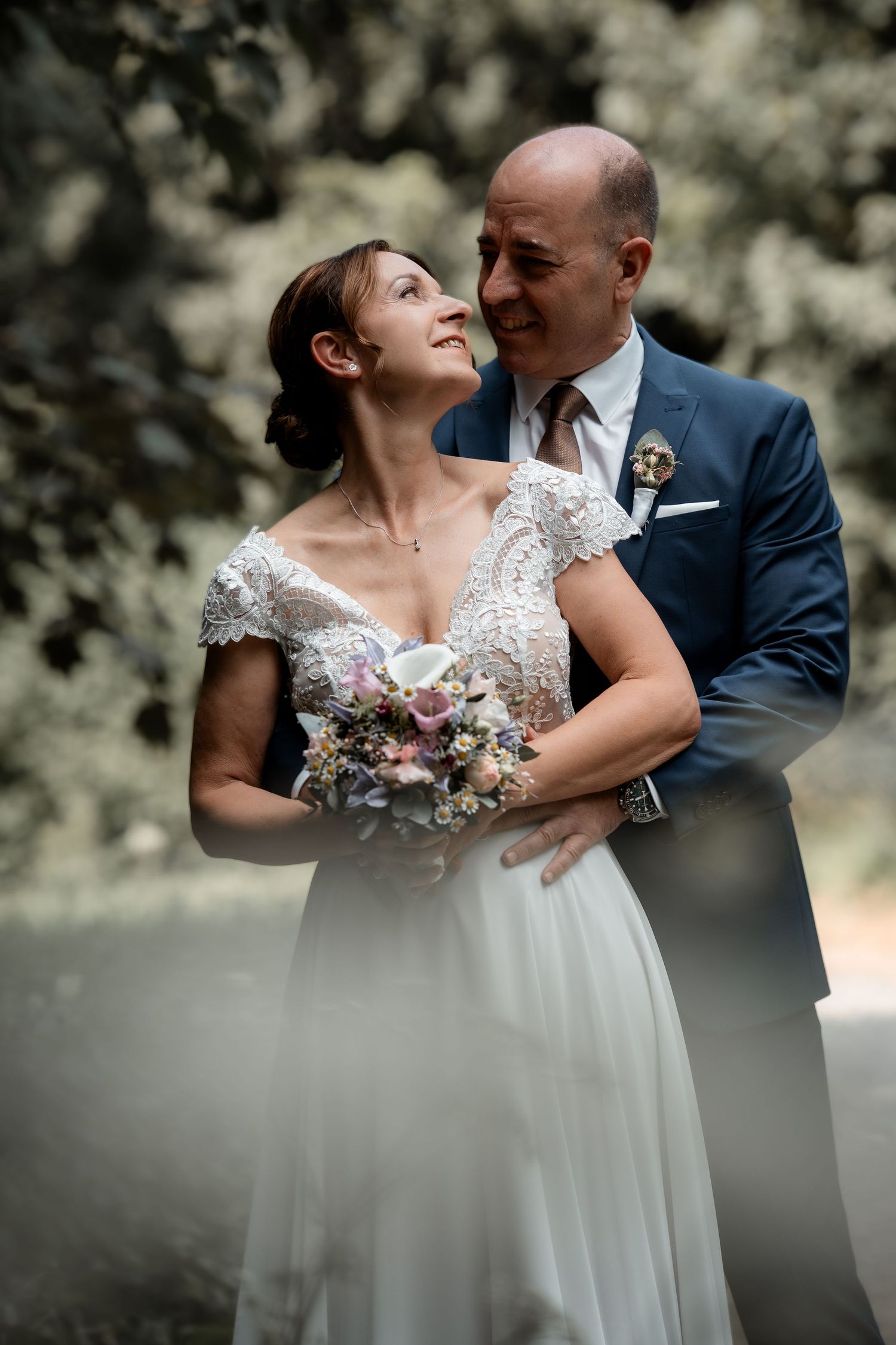 Bride and groom sharing a tender moment, smiling at each other, bride holding a pastel bouquet outdoors.