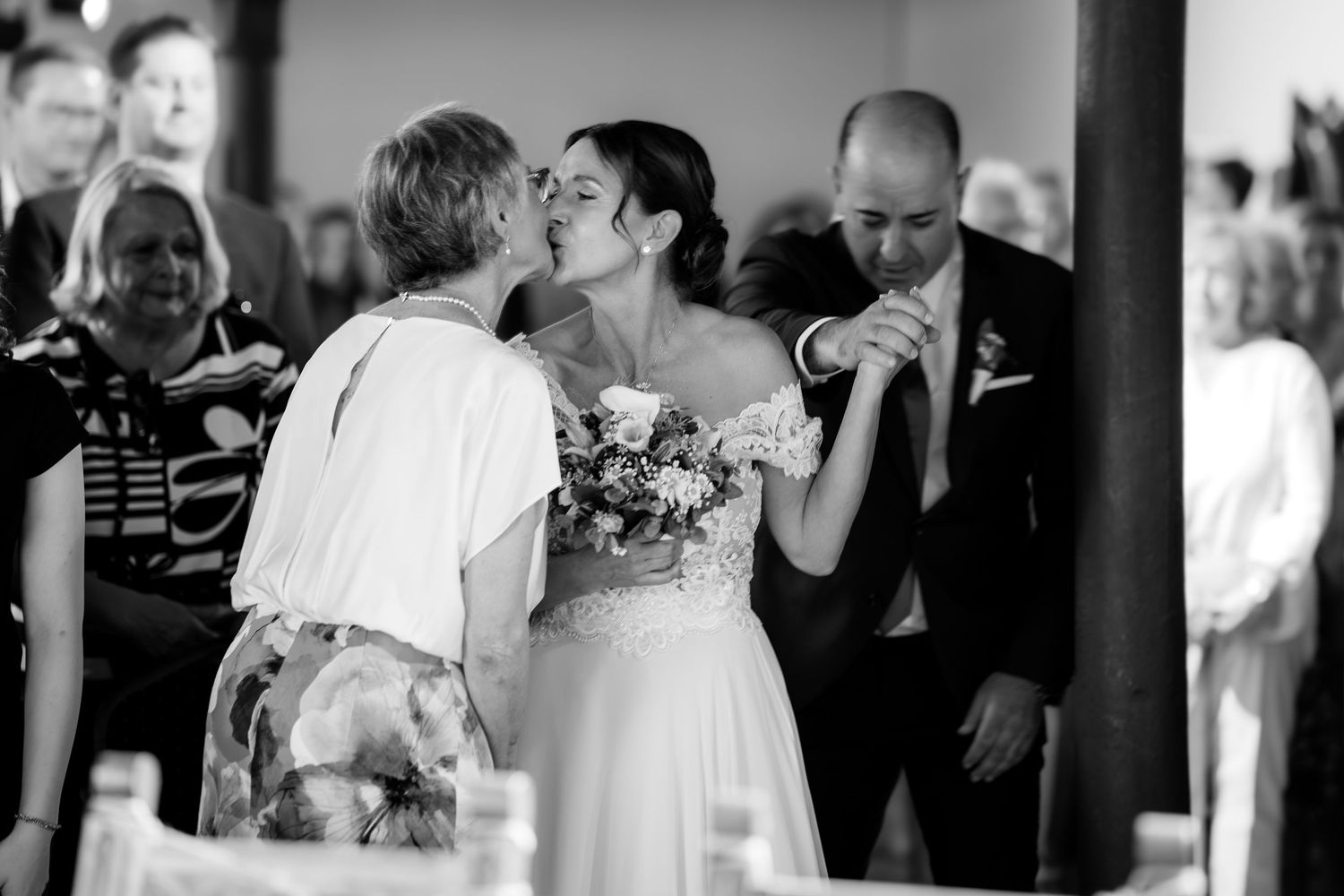 Black and white wedding photo of bride kissing guest while groom kisses hand of another guest indoors.