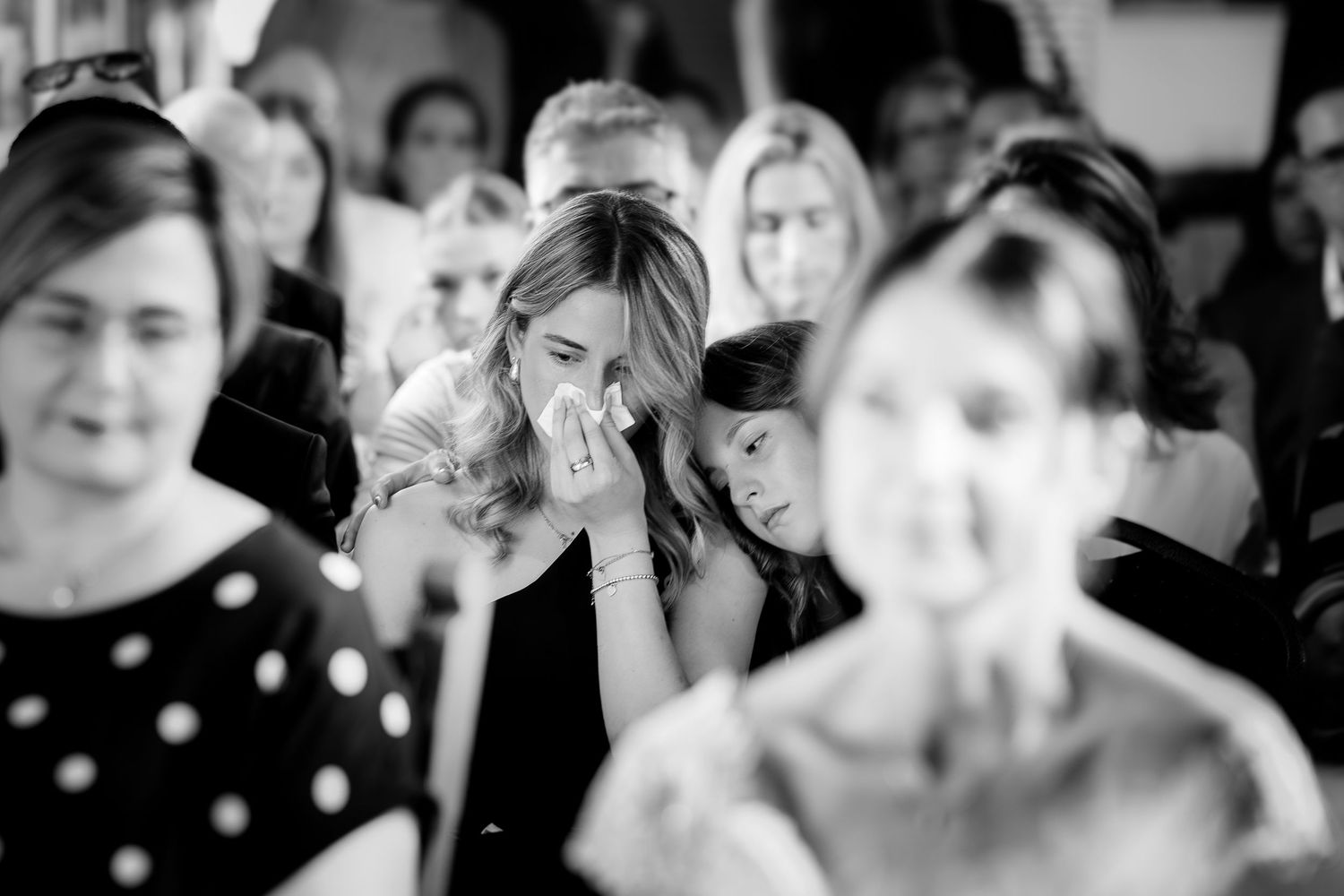 Black and white photo of emotional wedding guest wiping tears during ceremony surrounded by other guests.