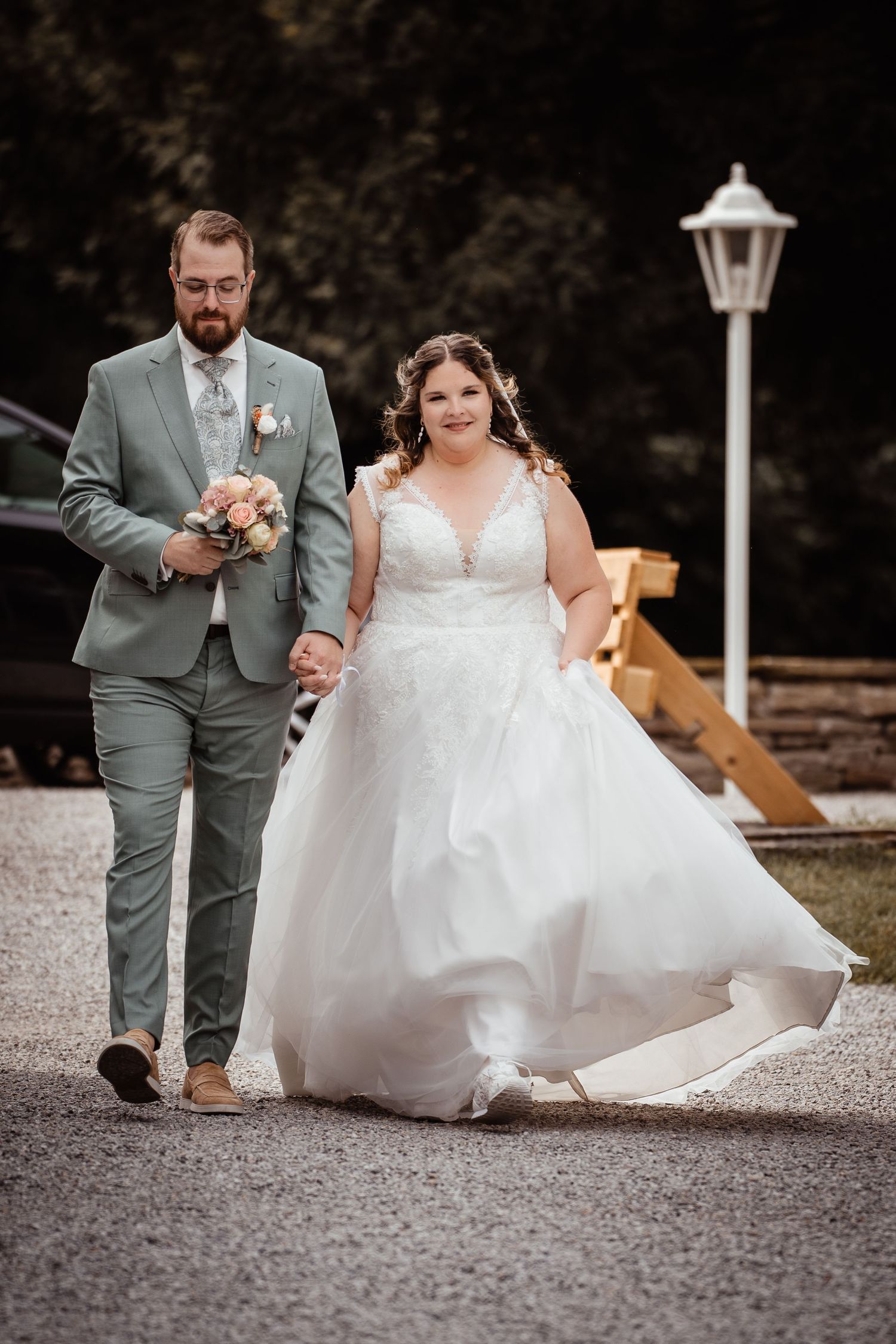 Bride in white ball gown and groom in sage green suit walk hand in hand on gravel path at outdoor wedding.