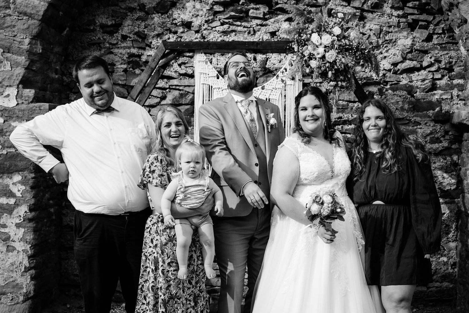 Wedding party of five adults and a baby pose together in black and white photo against a rustic stone wall.