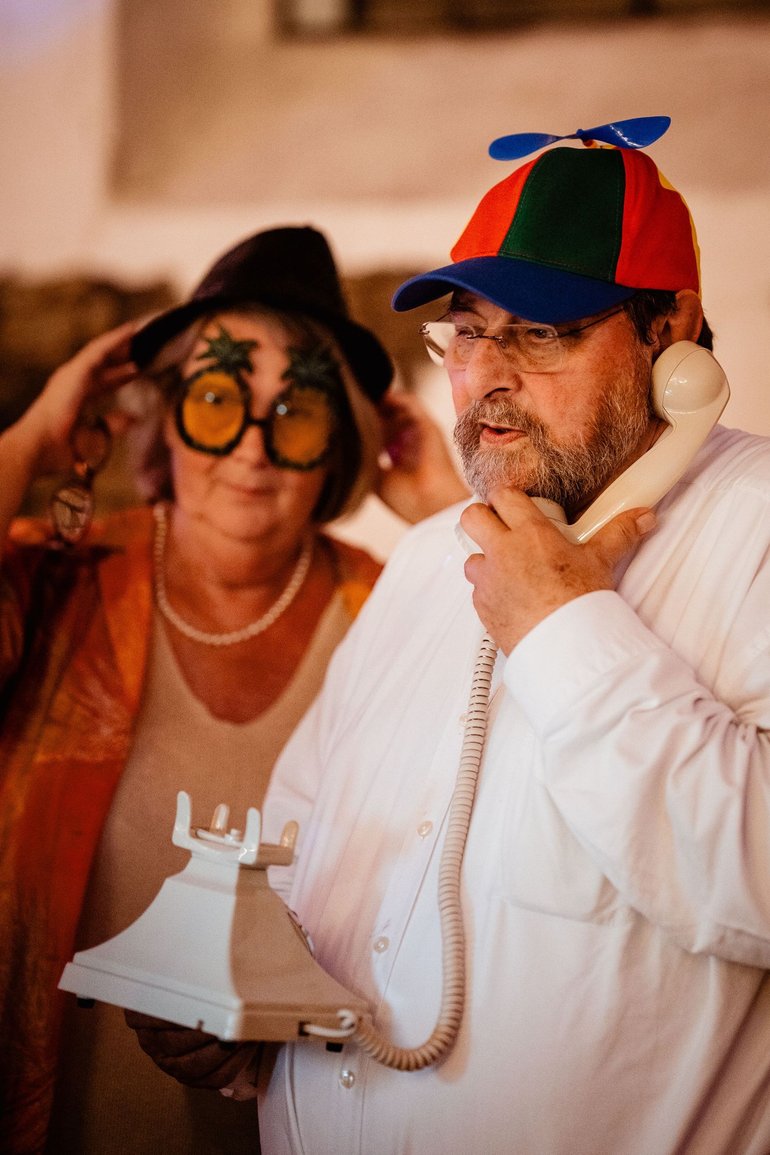 Man in colorful propeller hat speaks on vintage phone while woman wears novelty glasses at wedding.