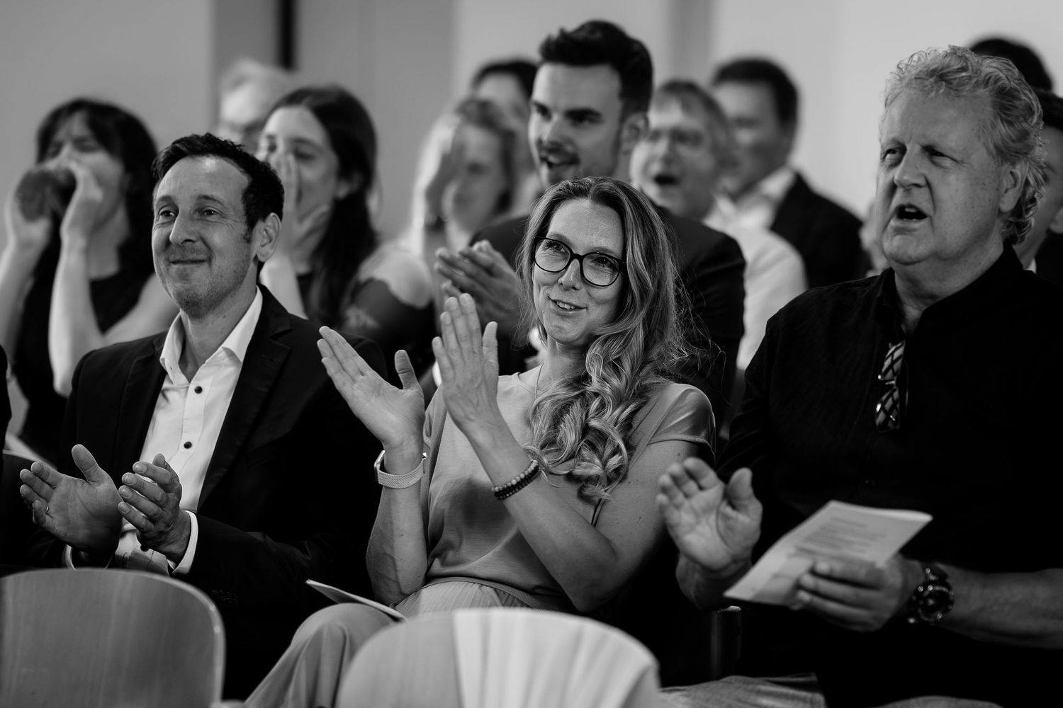 Wedding guests smile and applaud during ceremony, with woman in glasses and blonde hair at center.