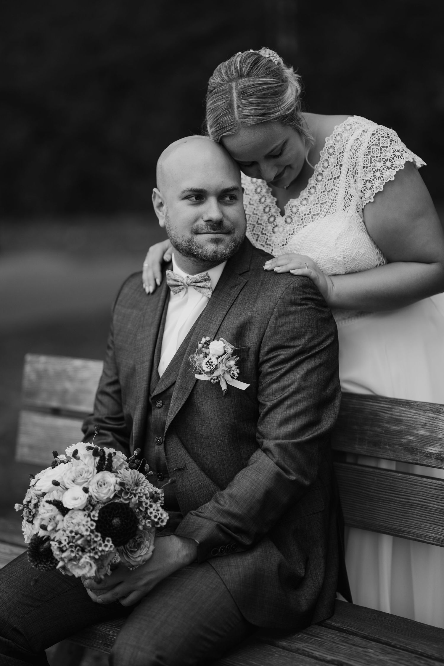Black and white wedding photo of a bald groom in a suit with a bride leaning on his shoulder, holding a bouquet.